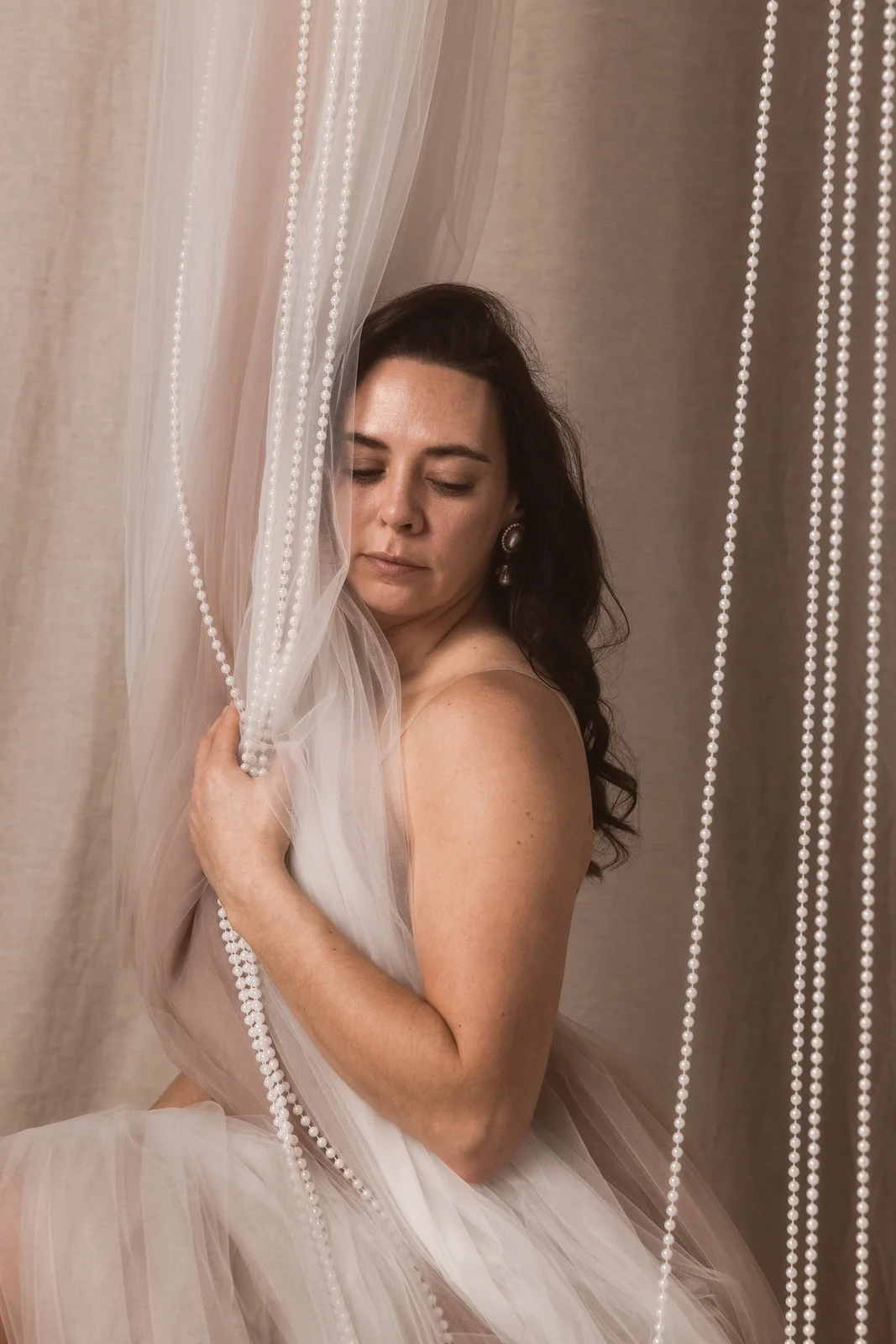 Buduarowy portret kobiety w klimacie filmowym. Kobieta trzyma zwisający z góry materiał i białe korale.