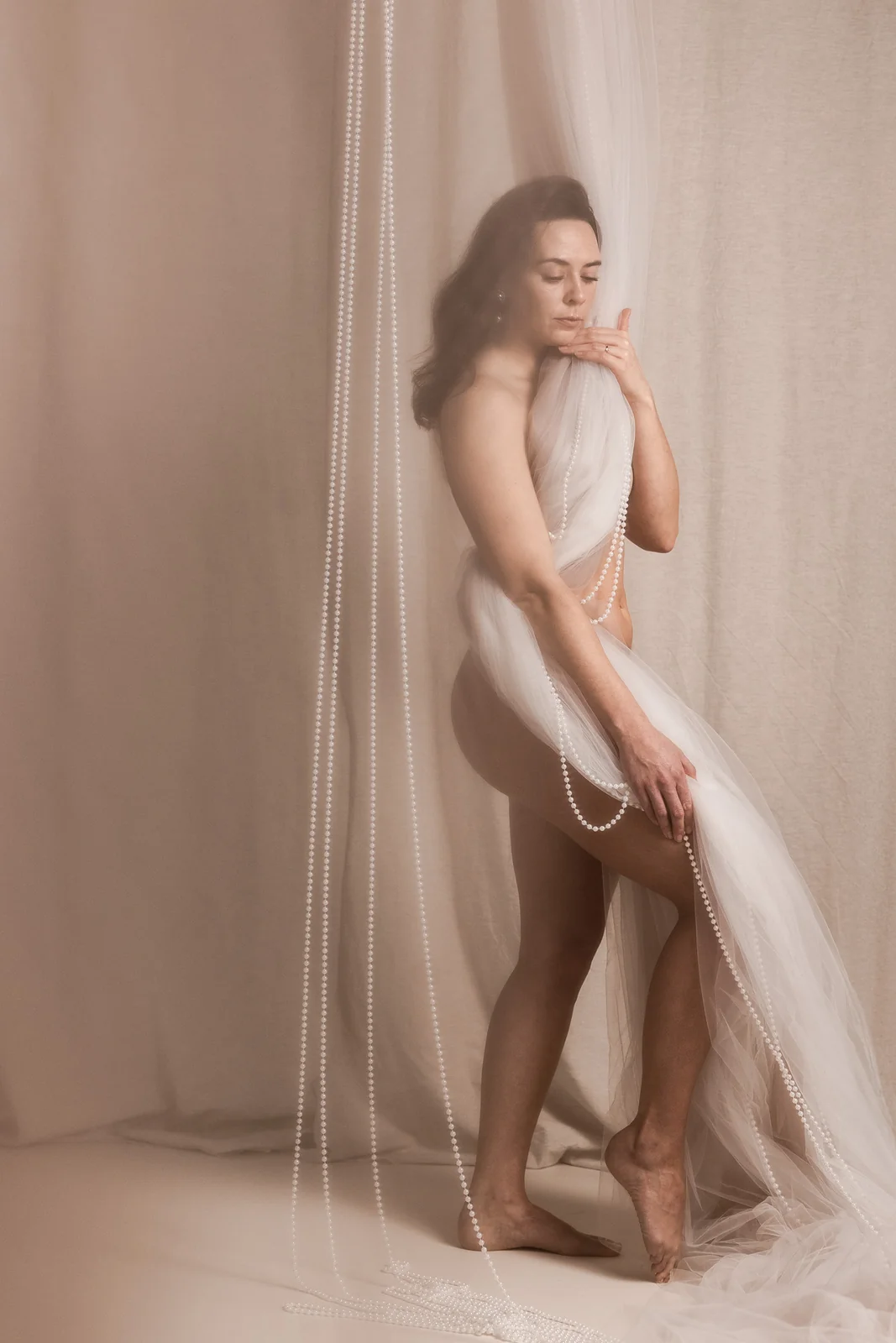 Buduarowa sesja kobieca w nastrojowym oświetleniu - kobieta zakryta miękkim jasnym tiulem