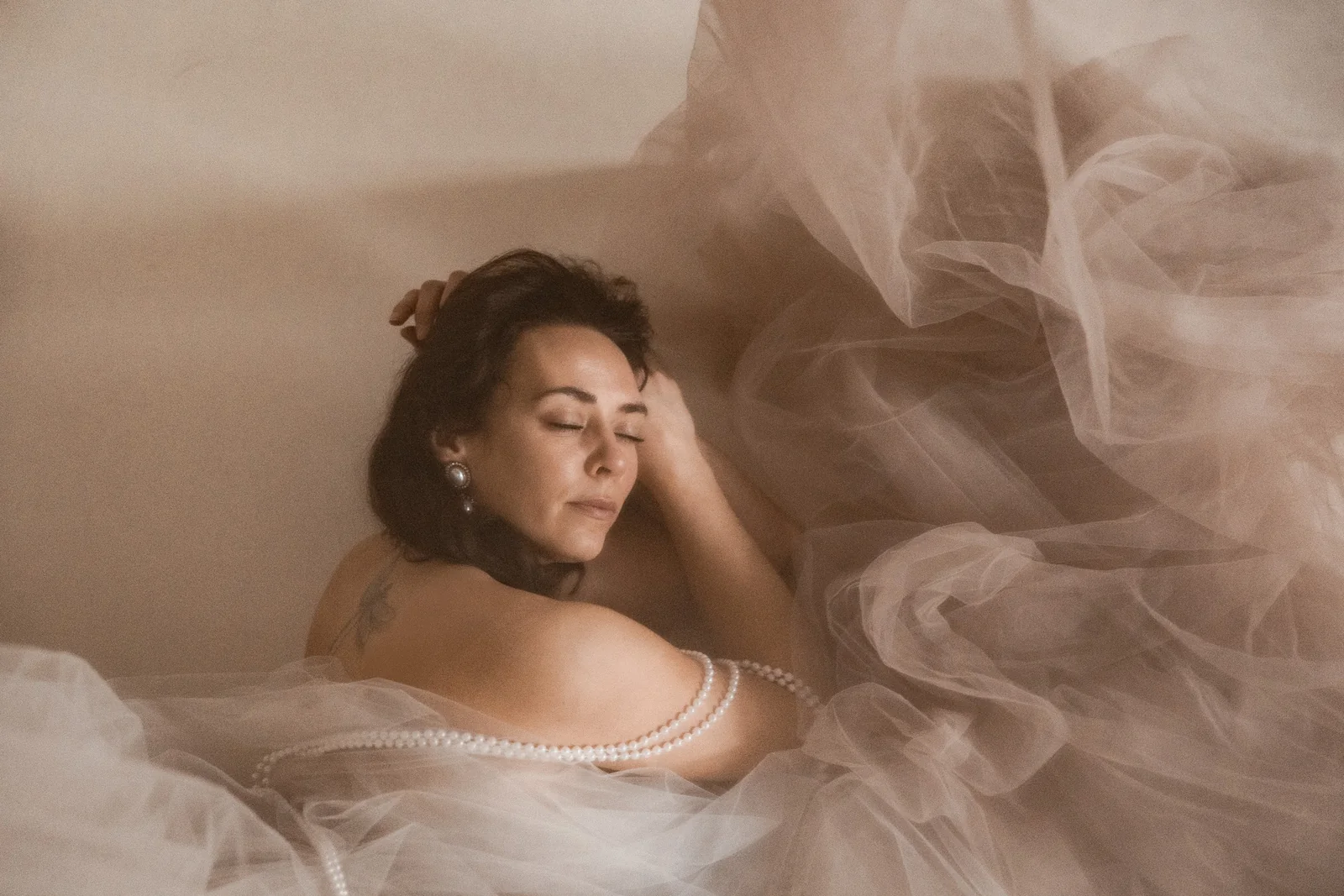 Kobieta w eleganckiej pozie z zamkniętymi oczami na tle rozproszonego minimalistycznego tiulu