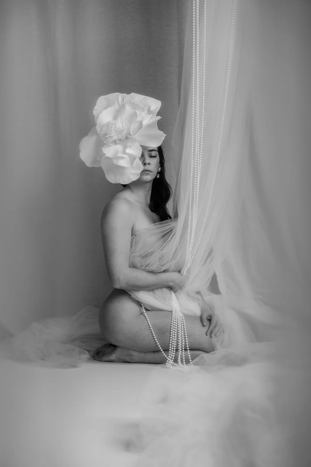 Czarno-biały portret kobiety w pozie siedzącej z duży białym kwiatem we włosach. Modelka otulona miękkim jasnym materiałem i koralami.