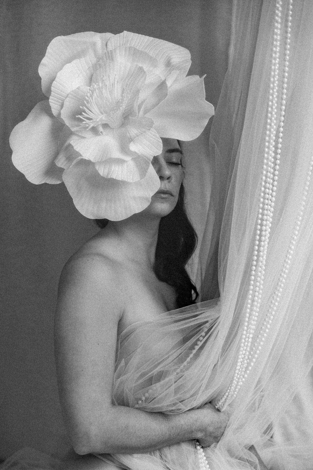 Portret kobiety w kontrastowym świetle i cieniu. Na pierwszym planie duży kwiat zdobiący włosy modelki, korale i tiule.