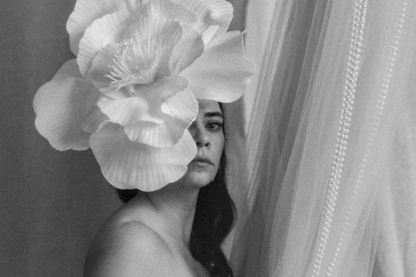 Czarno-biały portret kobiety, duży biały kwiat zdobi kobiece włosy. W tle tiule i białe korale.