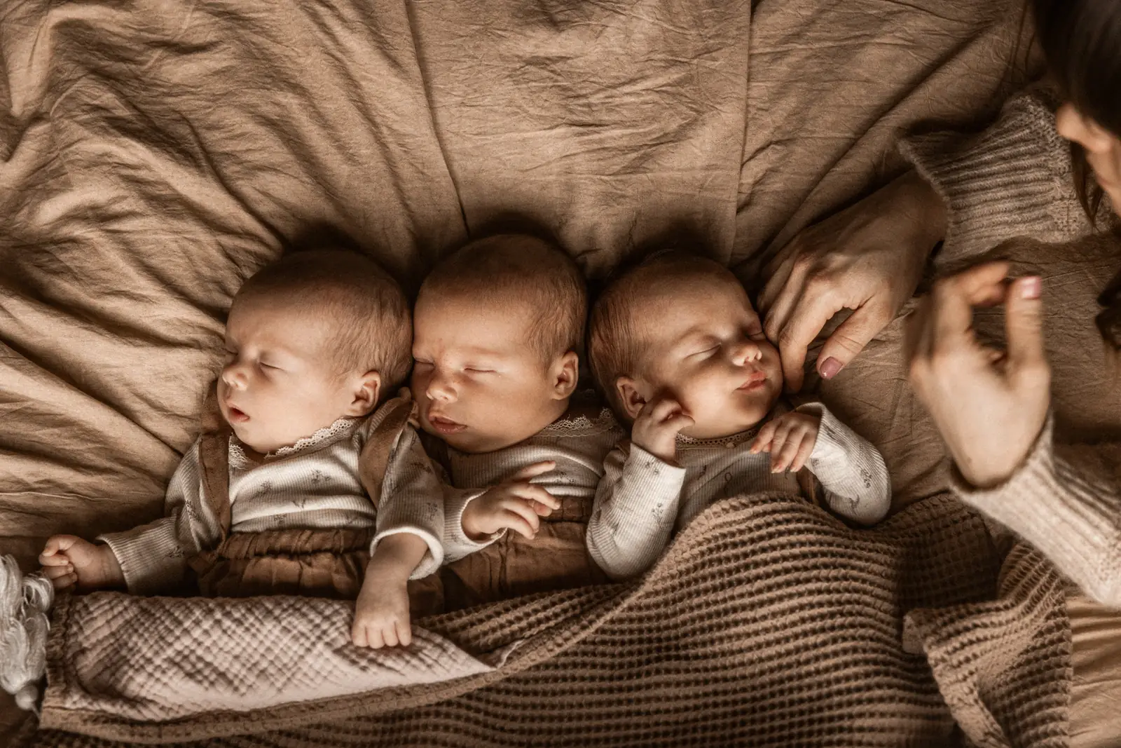 Trojaczki śpiące obok siebie na beanbag w domu podczas sesji noworodkowej, w tle dłonie mamy czule dotykające główkę dziecka