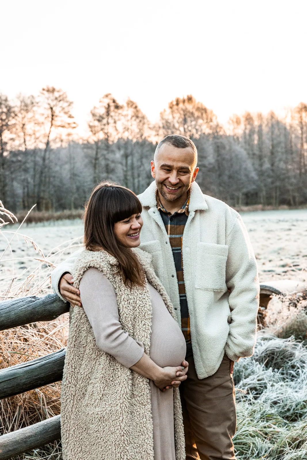 Przyszły tata obejmuje ukochaną partnerkę, a przyszła mama obejmuje rękami brzuszek. W tle wiejski krajobraz i szron na łące.