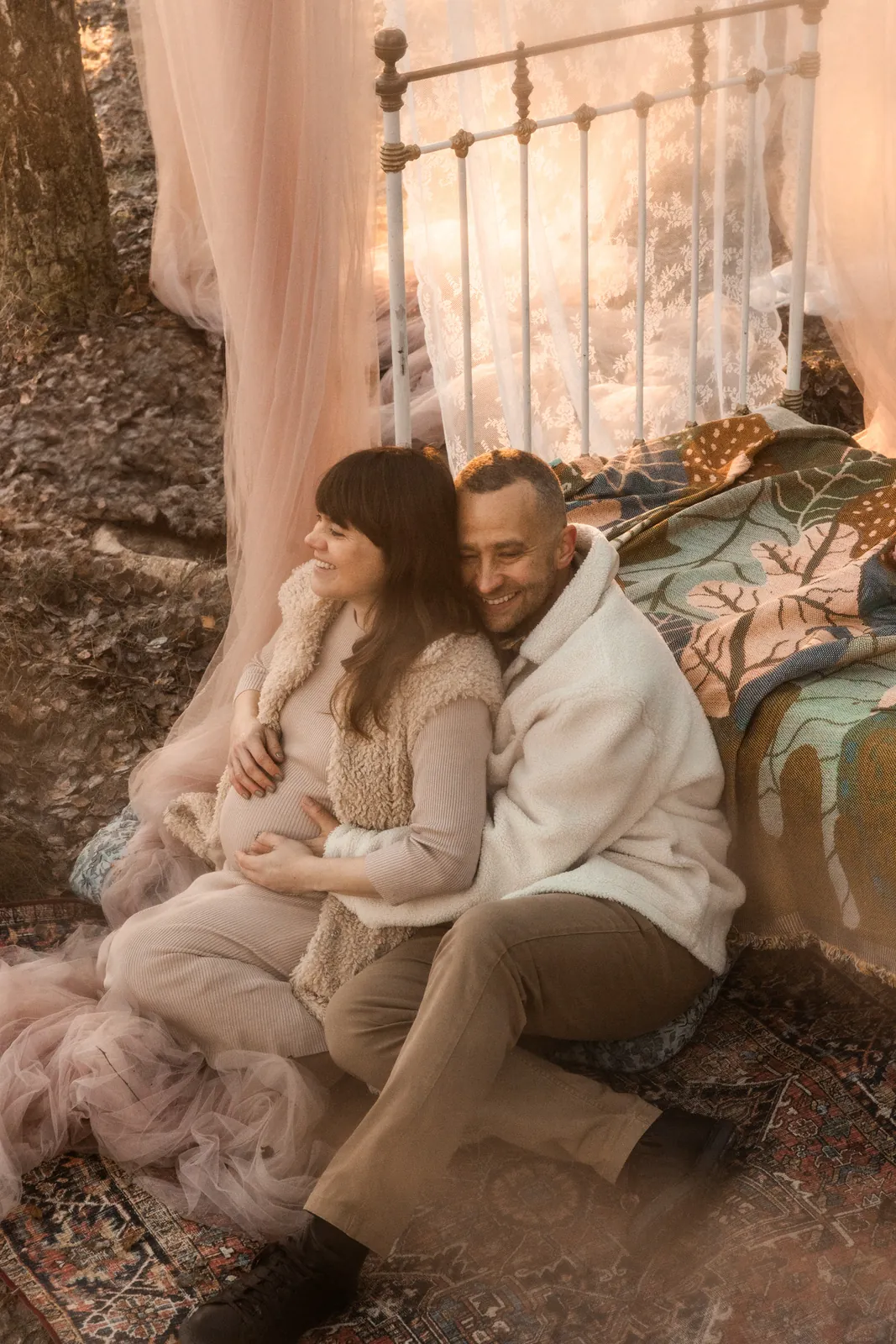 Rodzice przytuleni do siebie uśmiechają się podczas sesji ciążowej w lesie. Para oparta o stylizowane łóżko w środku lasu.