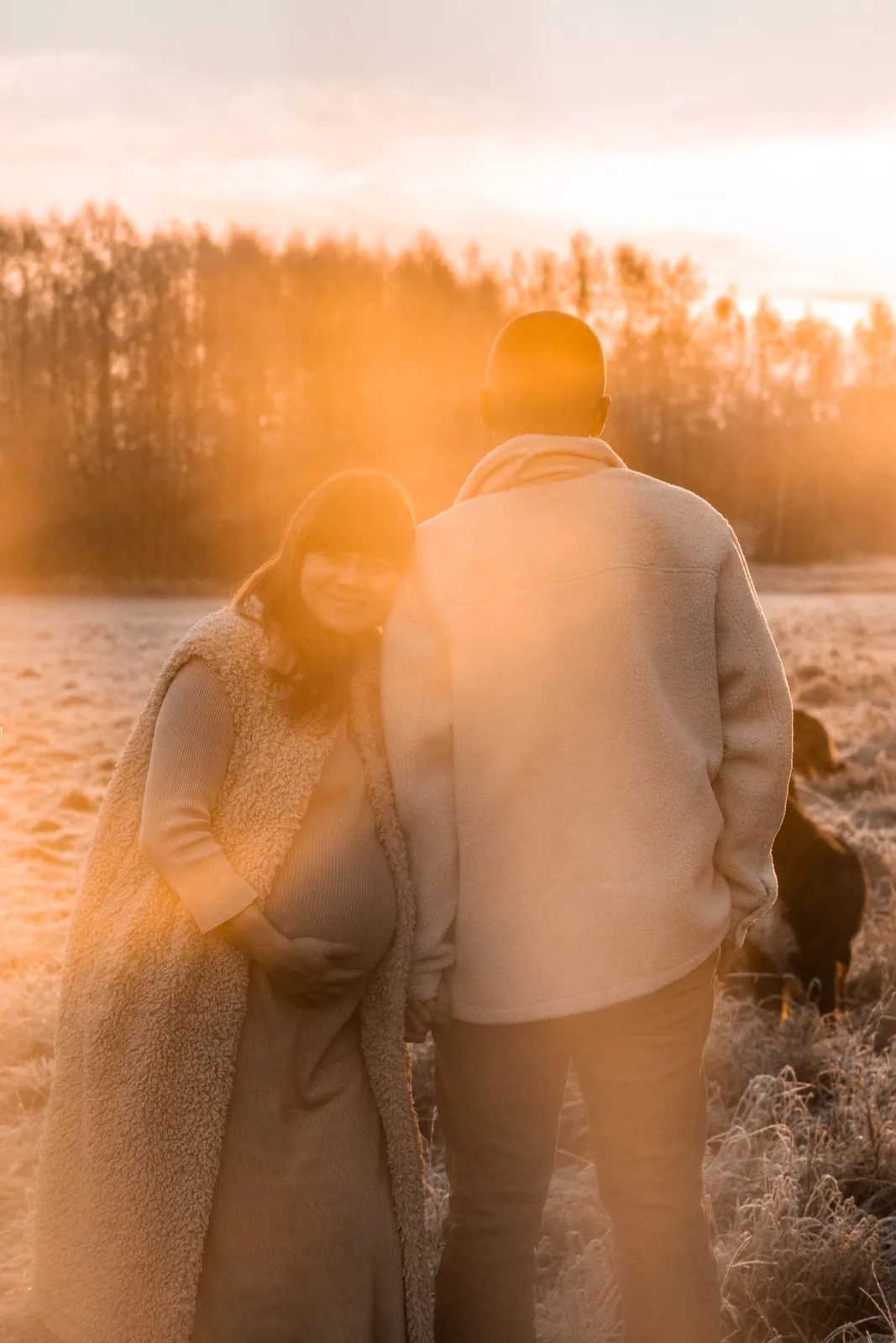 Kobieta w ciąży przytulona do partnera na tle oszronionej łąki i jesiennych drzew