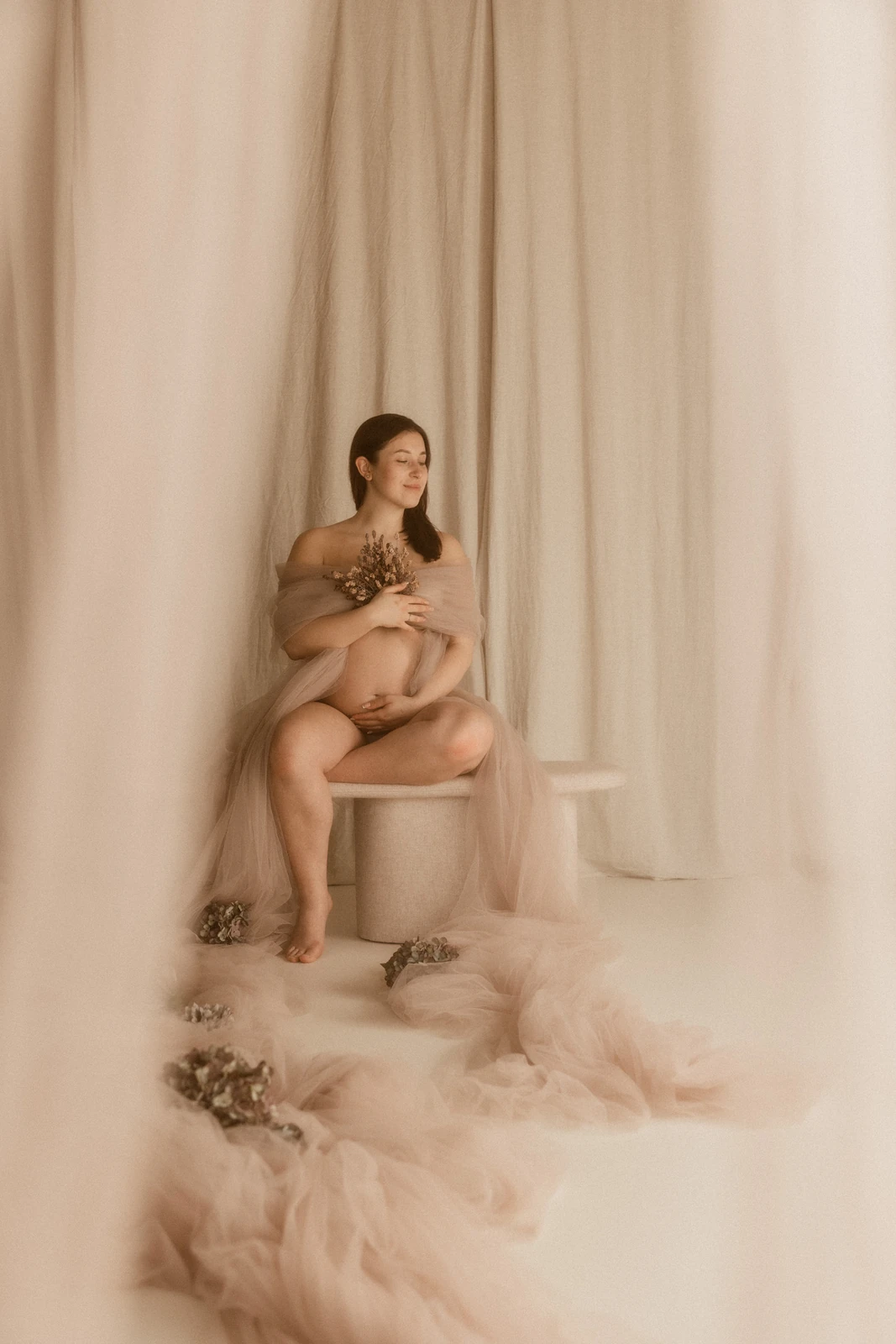 Sensualna sesja ciążowa w studiu – kobieta z kwiatami w delikatnym świetle otulona tkaniną