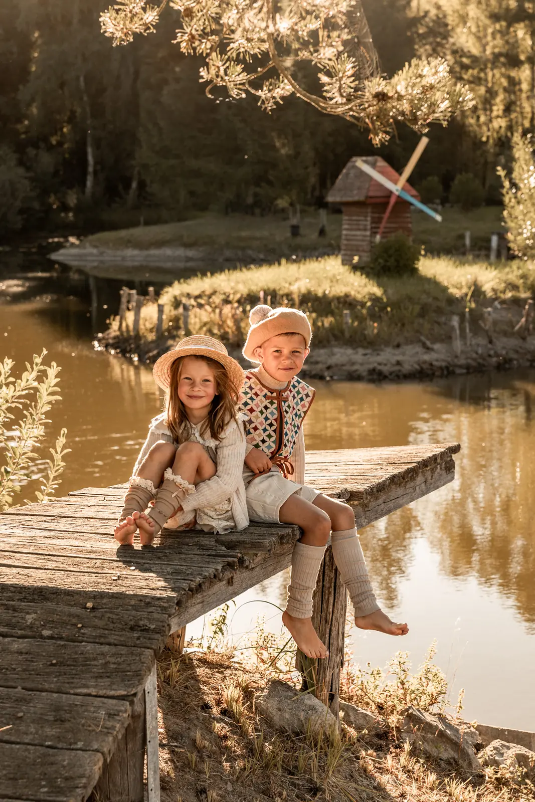 Brat z siostrą w magicznej scenerii plenerowej, siedzą na drewnianym mostku i uśmiechając się spoglądają w obiektyw