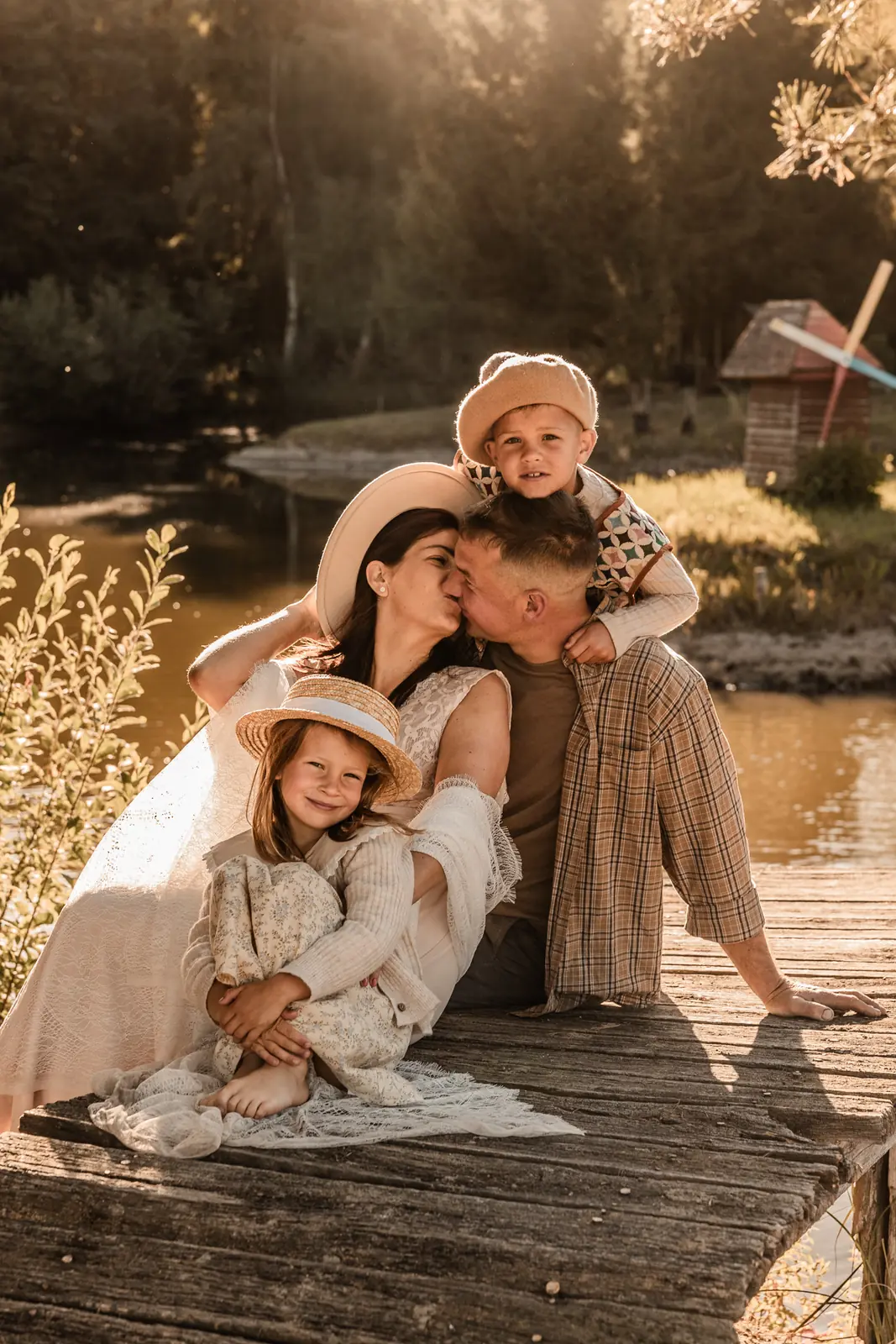 Rodzice z dziećmi siedzą na mostku nad jeziorem przytuleni do siebie w naturalnym kadrze rodzinnej sesji w plenerze