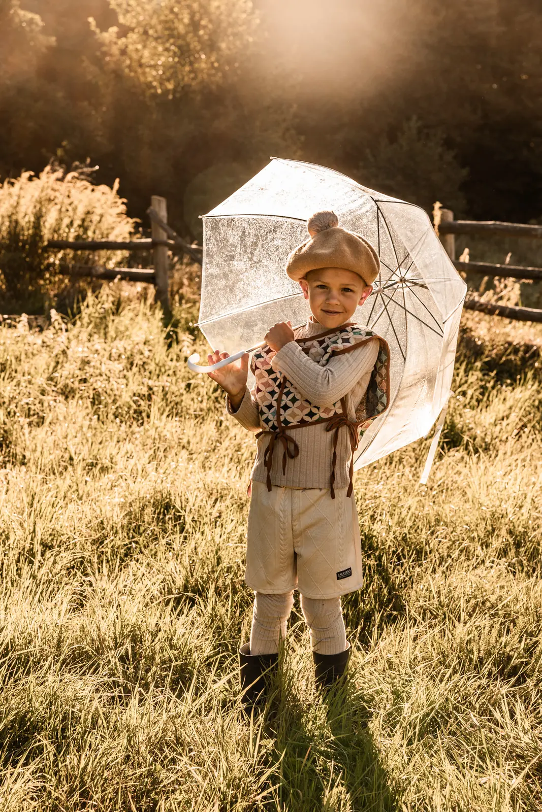 Chłopiec z parasolem podczas zabawy na łące w plenerze. W tle sielankowy krajobraz o wschodzie słońca.