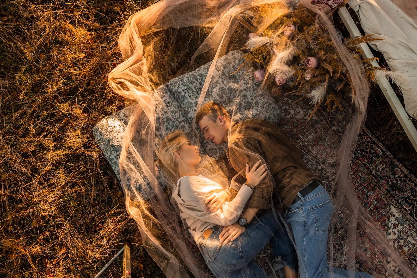 Para leży na stylizowanym dywanie przykryta przezroczystym materiałem, który rozszczepia promienie słoneczne podczas sesji narzeczeńskiej