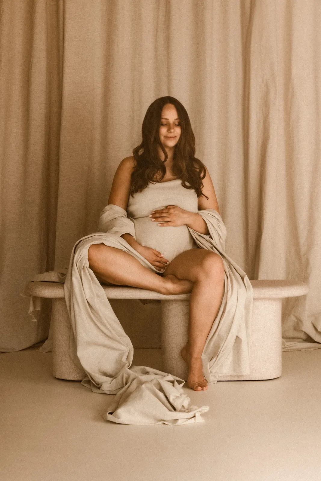 Kobieta z zamkniętymi oczami siedzi na ławce ubrana w jasne tkaniny. Dotyka czule swój brzuszek podczas sesji w studiu.