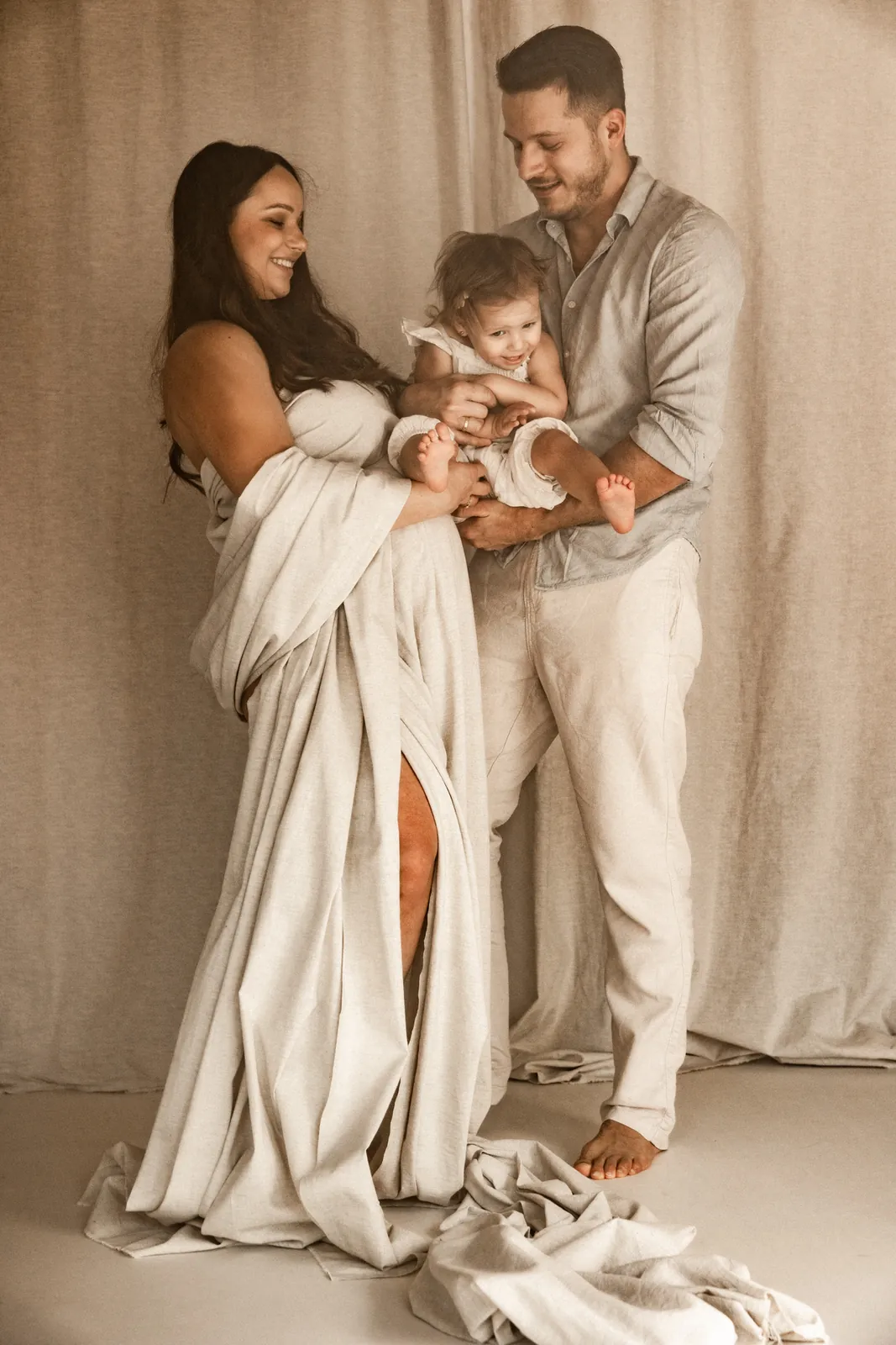 Przyszli rodzice trzymają wspólnie starsze dziecko na rękach w trakcie sesji rodzinnej sesji ciążowej