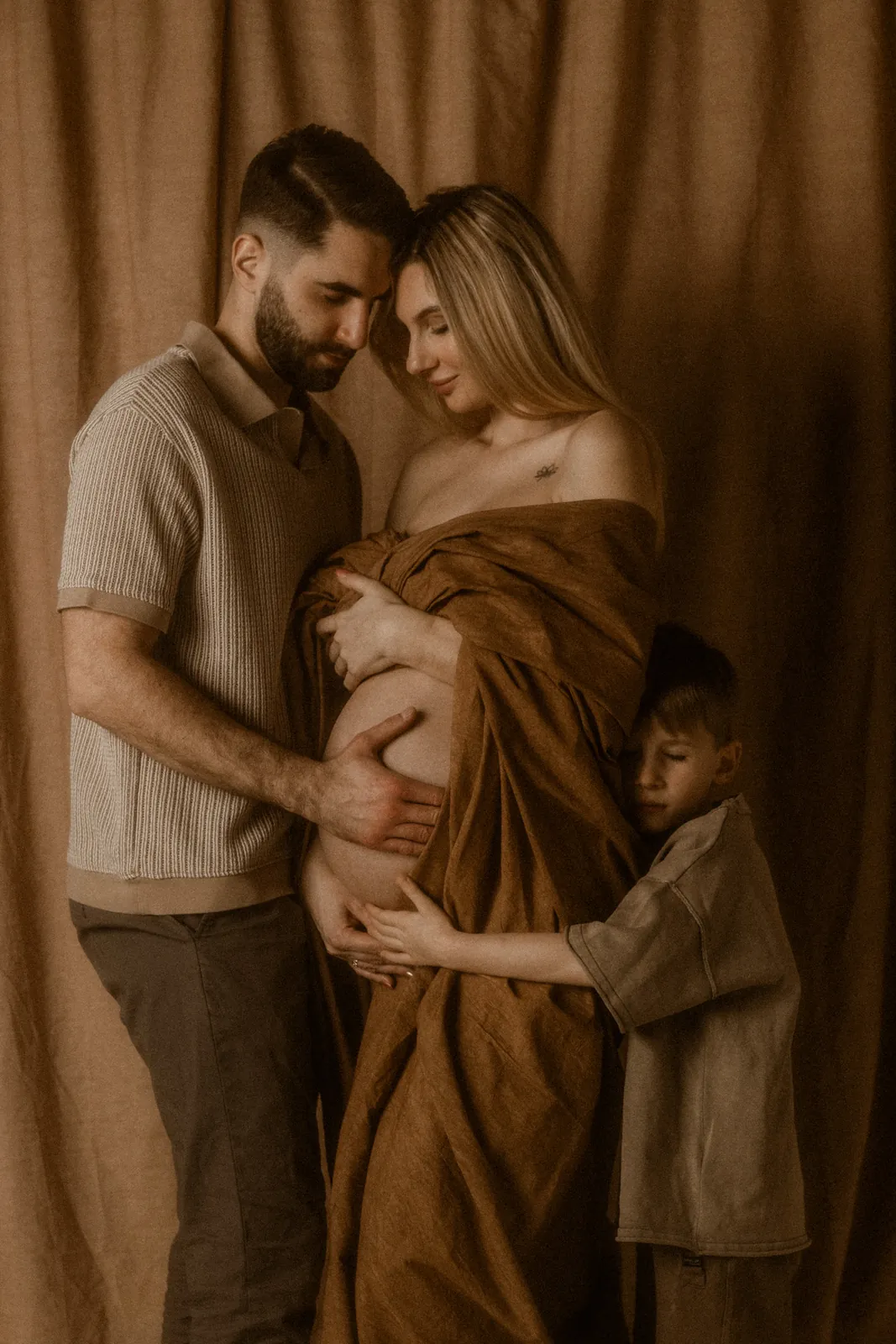 Przyszła mama z rodziną podczas spokojnej sesji ciążowej w studio. Tata i starsze dziecko przytulają brzuszek mamy.