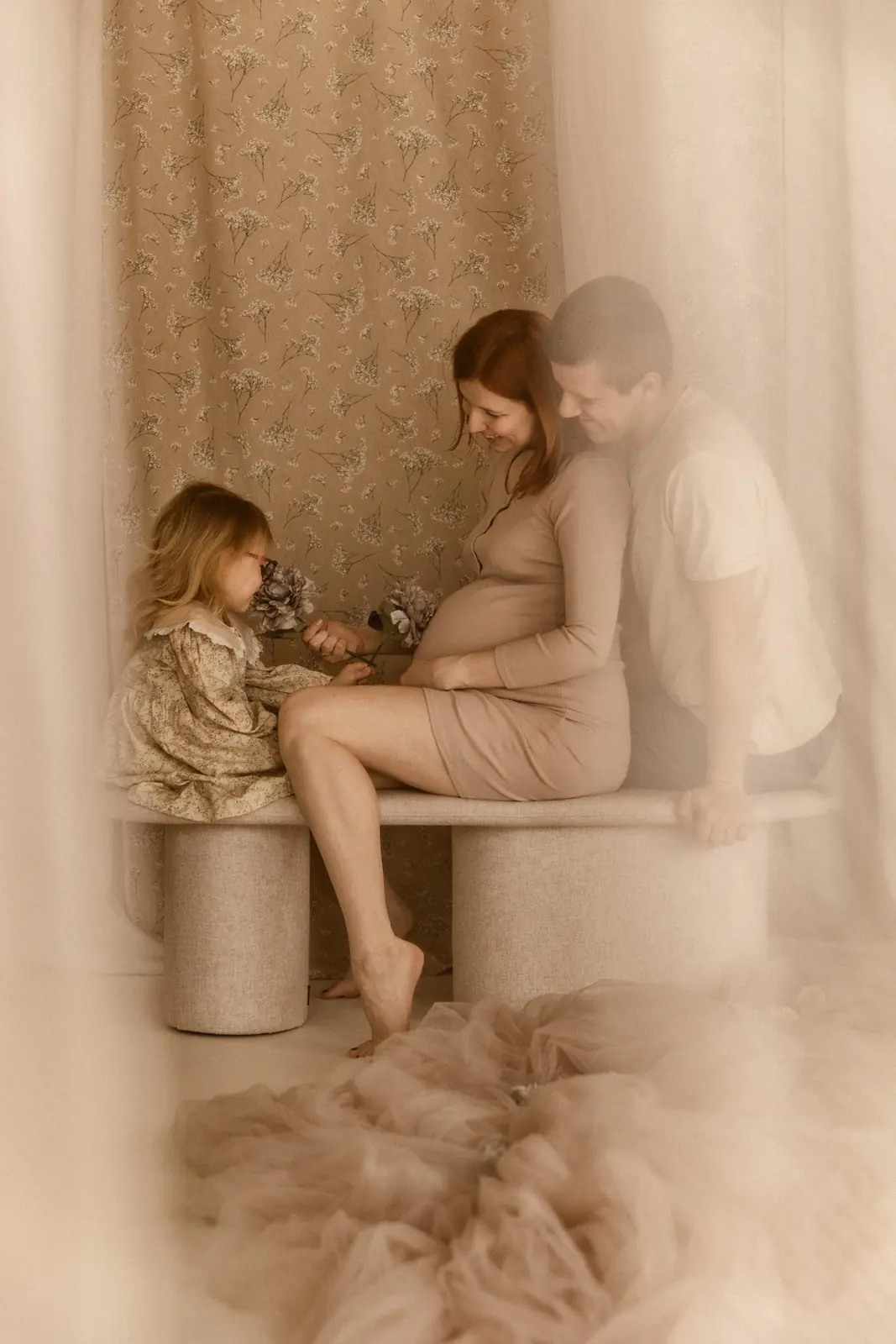 Przyszli rodzice spędzają mile czas z starszym dzieckiem siedząc na ławce, trzymając w ręce kwiaty, w tle stylizowane beżowe tło.