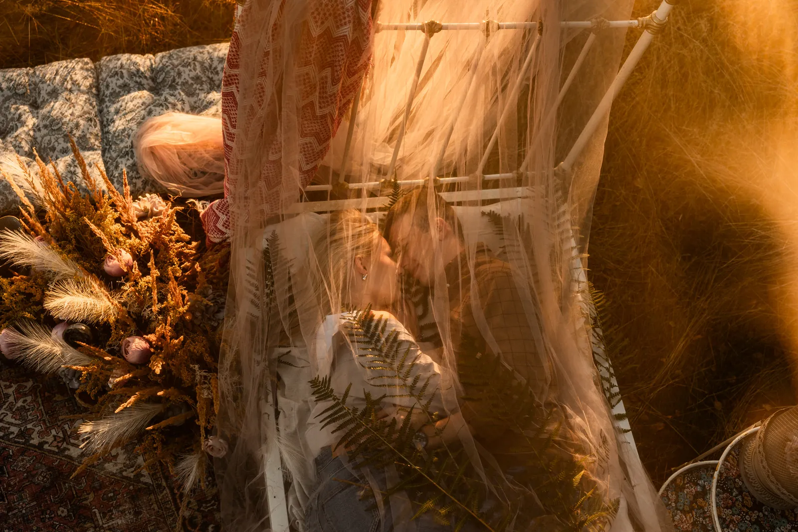 Zakochana para podczas plenerowej sesji zdjęciowej leży na łóżku pośrodku łąki, otula ich delikatny materiał, w tle naturalna kompozycja