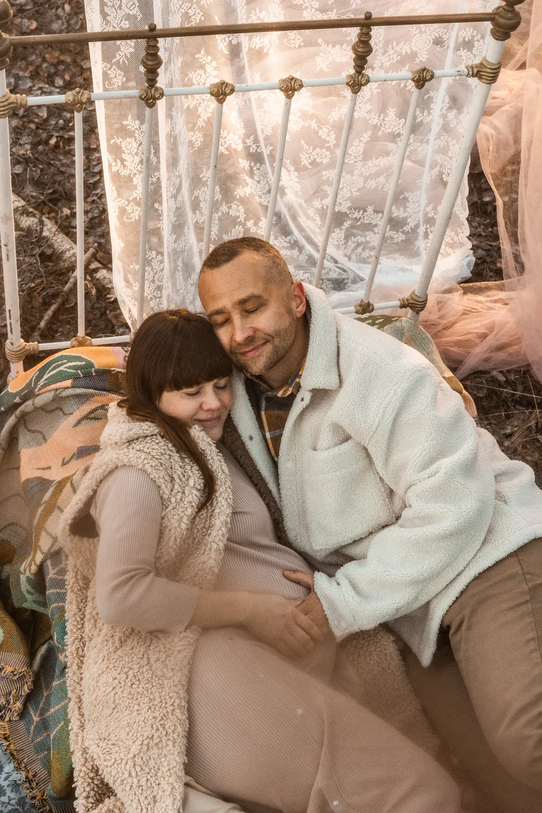 Para leży przytulona na zdobionym łóżku w środku lasu podczas sesji brzuszkowej w plenerze