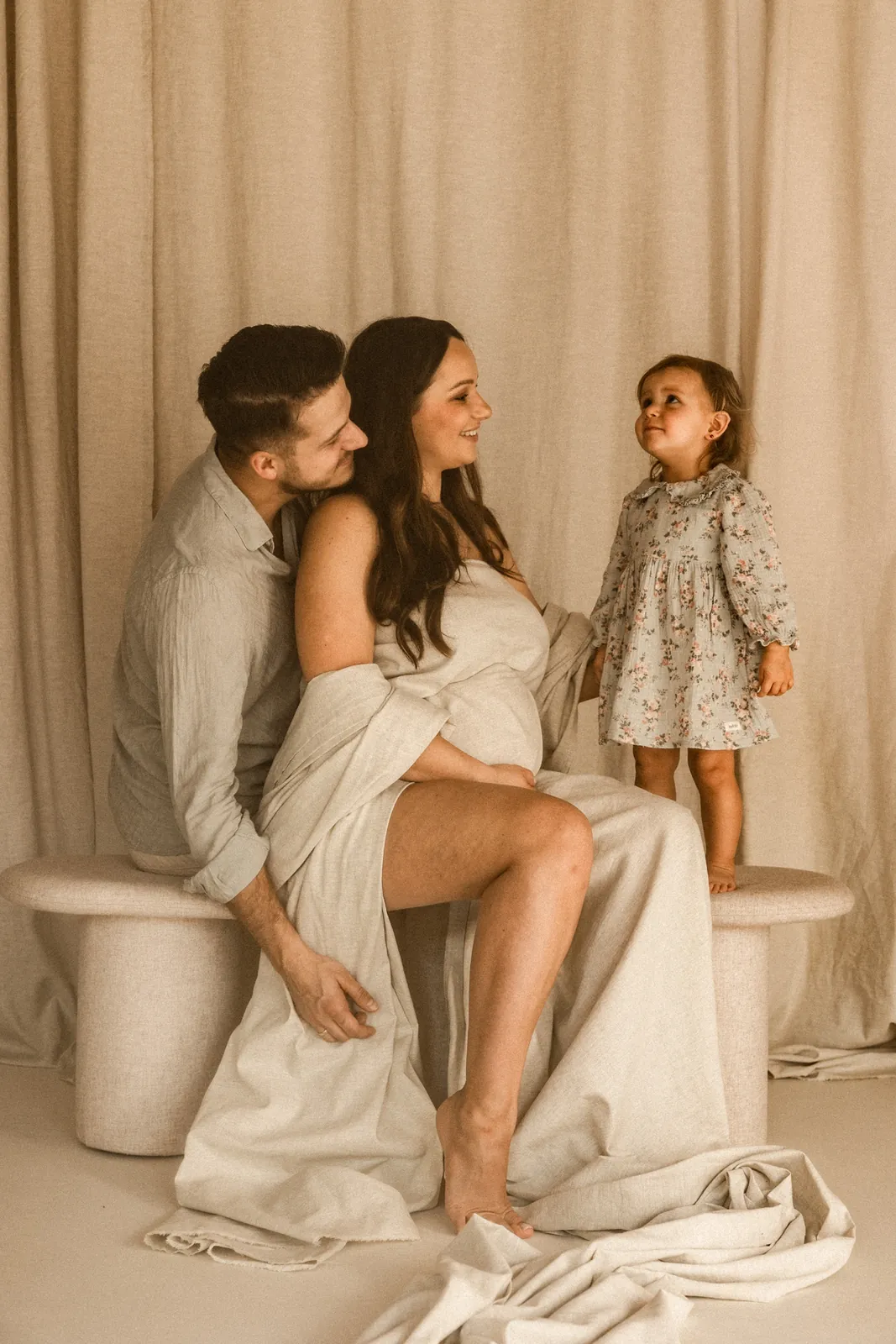 Córeczka stoi na jasnej ławce obok siedzących rodziców, w tle kremowe tło z delikatnych materiałów