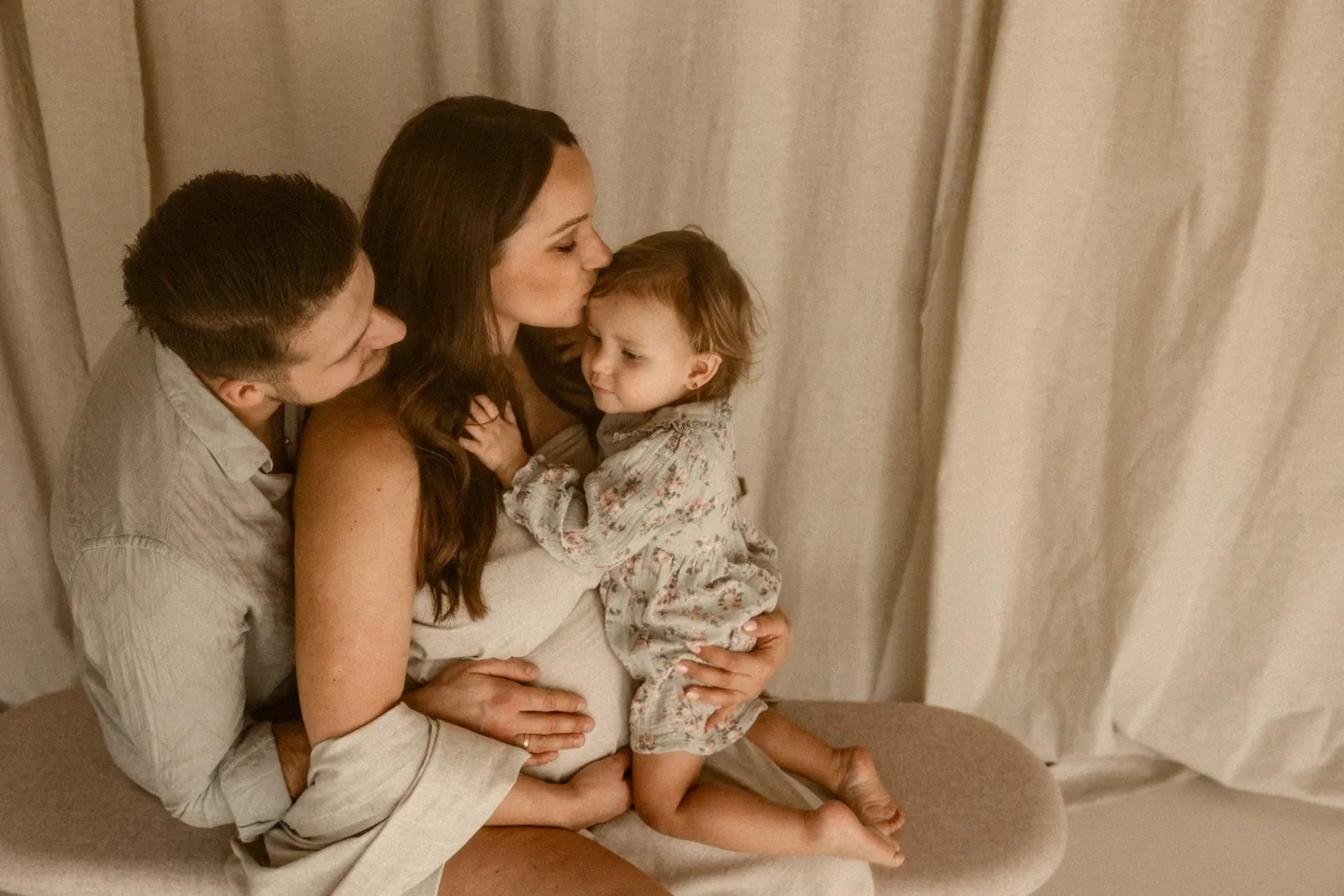 Przyszła mama całuje czule w główkę starszego dziecka, tata przygląda się przytulając brzuszek mamy