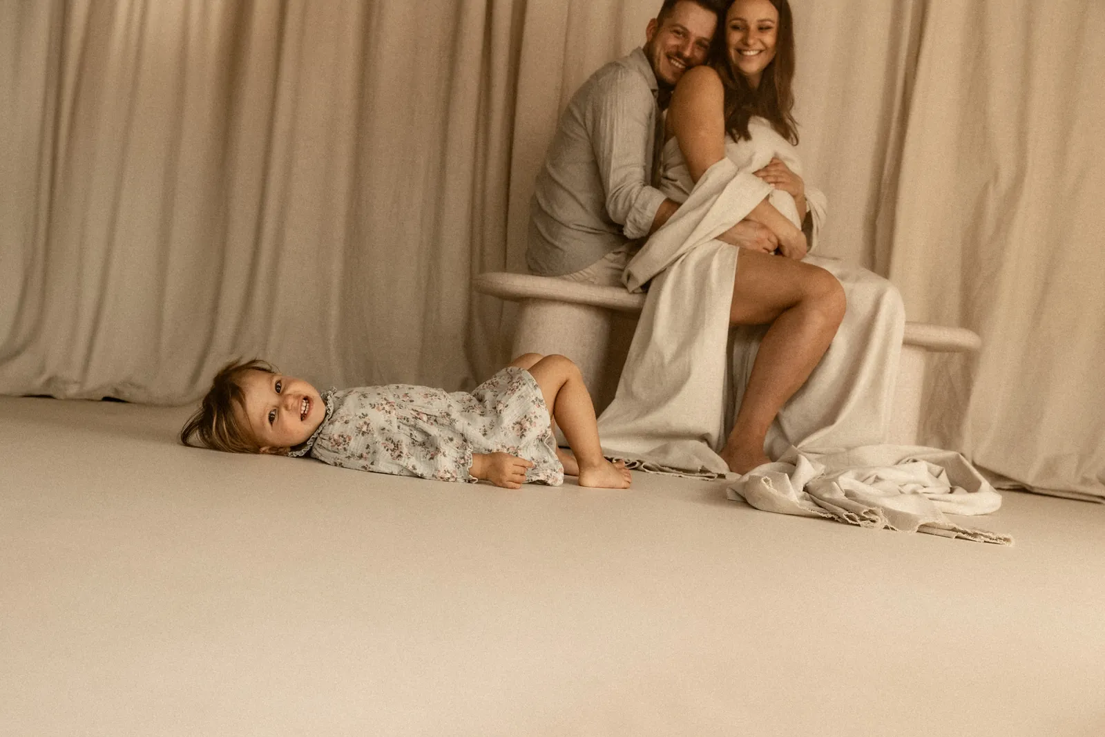 Rodzinne zdjęcie w stylu filmowym - dziecko leży wesoło na podłodze, a rodzice za nim na ławce śmieją się głośno