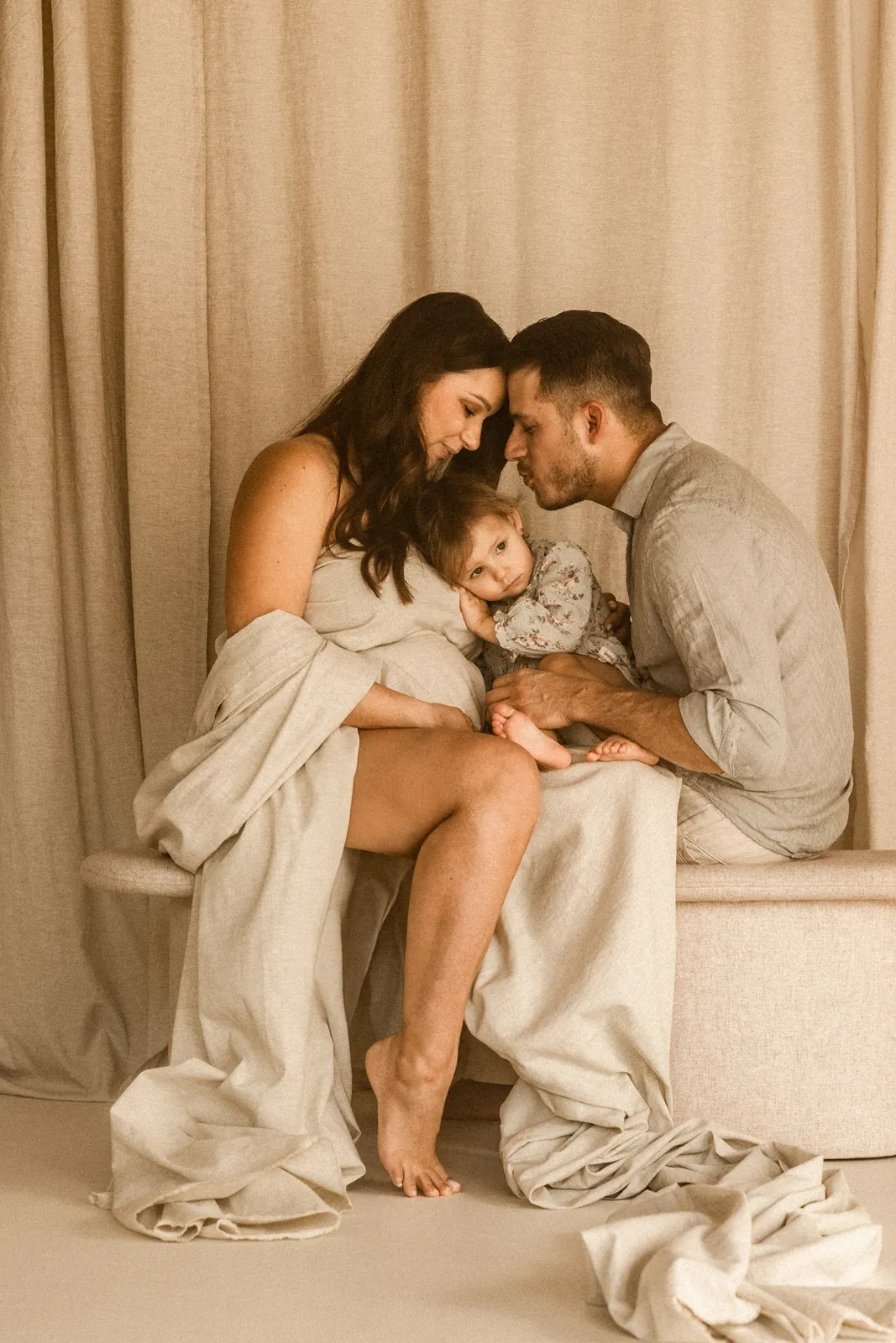 Rodzinne zdjęcie przyszłych rodziców i ich córki podczas minimalistycznej sesji ciążowej w studiu. Dziecko przytulone do mamy brzuszka.