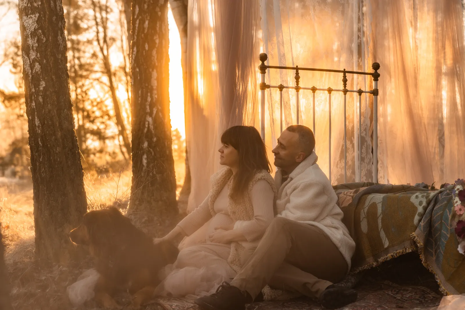 Fotografia kobiety w ciąży w naturalnym świetle. Przyszła mama czule objęta przez partnera, w towarzystwie ich pupila. W tle las i piękny wschód słońca.