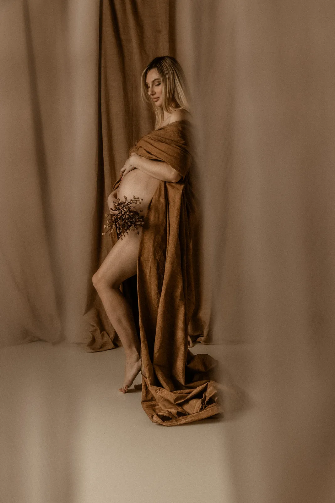 Zmysłowe portret kobiety w ciąży w minimalistycznym studiu. Przyszła mama owinięta w brązową tkaninę z kwiatem w ręce.