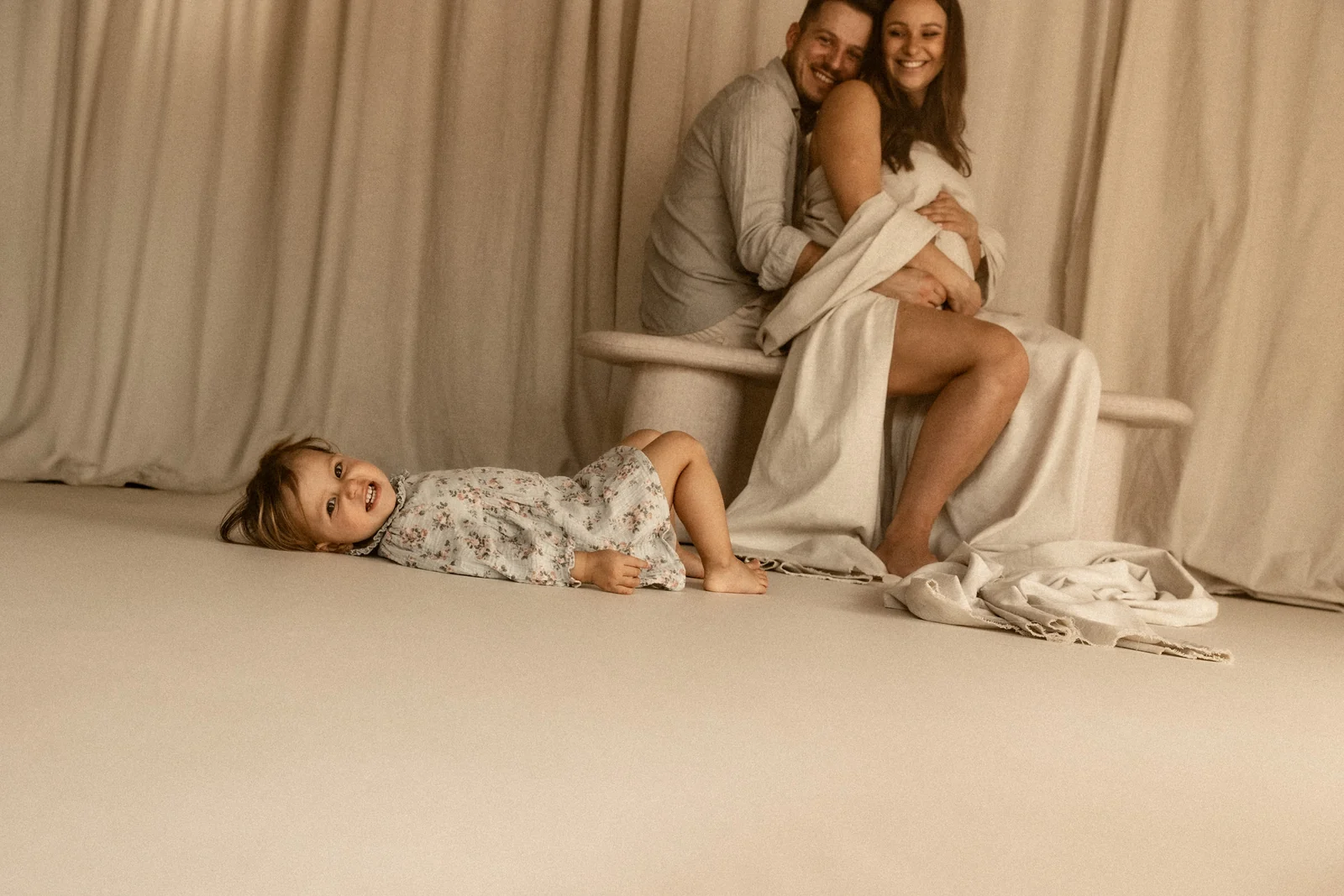 Rodzinne ujęcie w stylu filmowym na którym rodzice siedzą na ławce, a córeczka leży obok rodziców na białej podłodze. Tata przytula delikatnie brzuch przyszłej mamy.