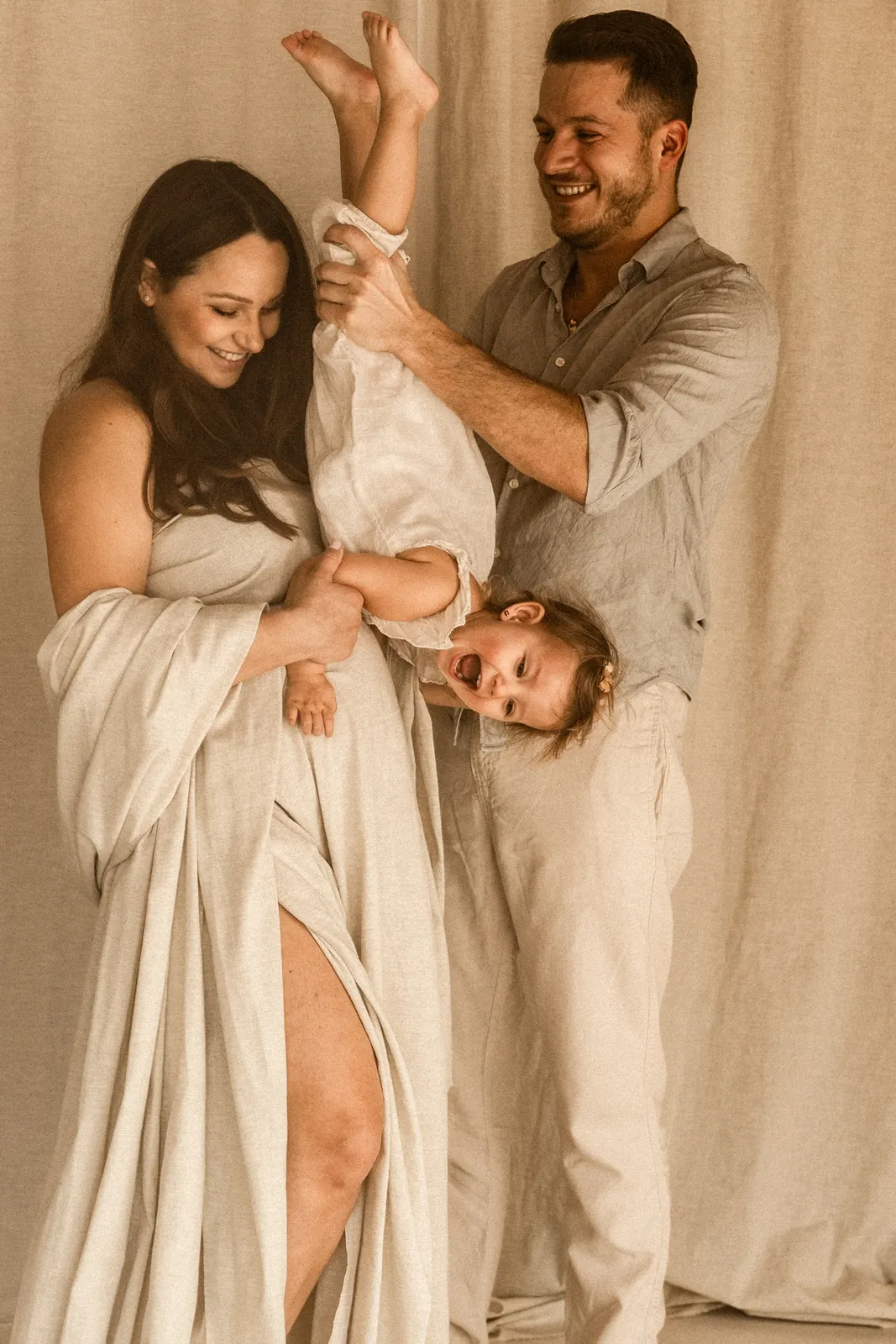 Emocjonalne i zabawne ujęcie taty trzymającego za nogi swoją córkę, która radośnie próbuje przytulić brzuszek swojej mamy.