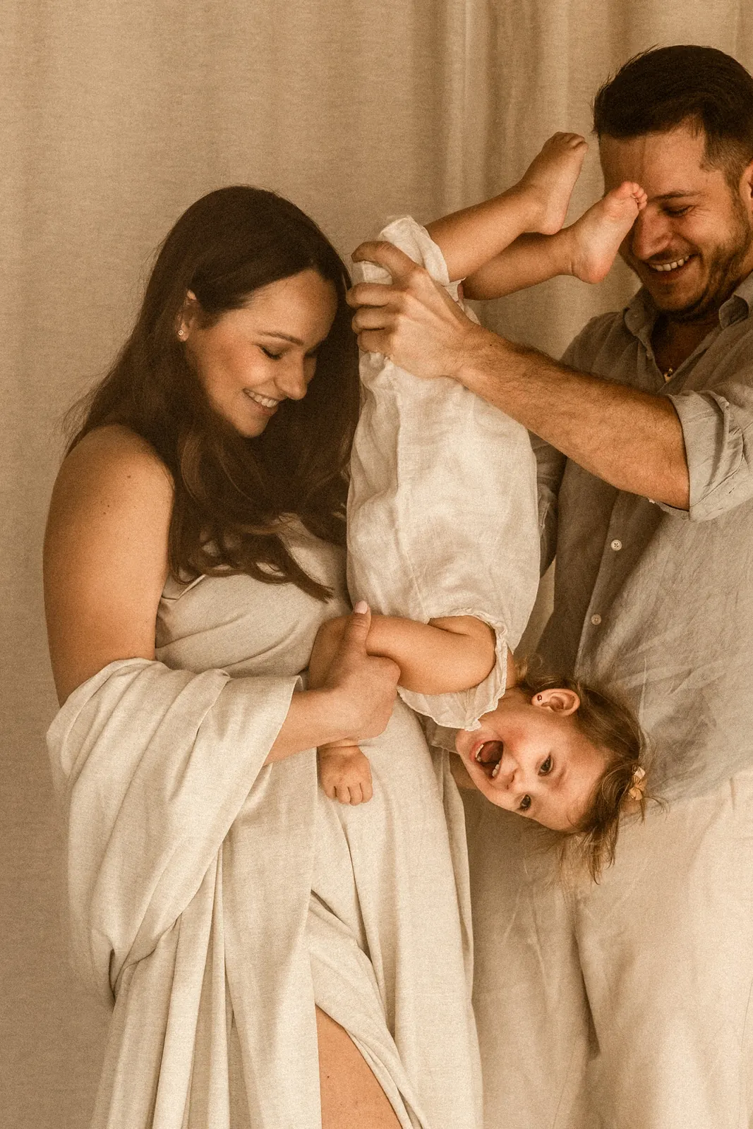 Zabawny moment rodzinnej sesji ciążowej, tata trzyma dziecko do góry nogami, mama obserwuje scenę
