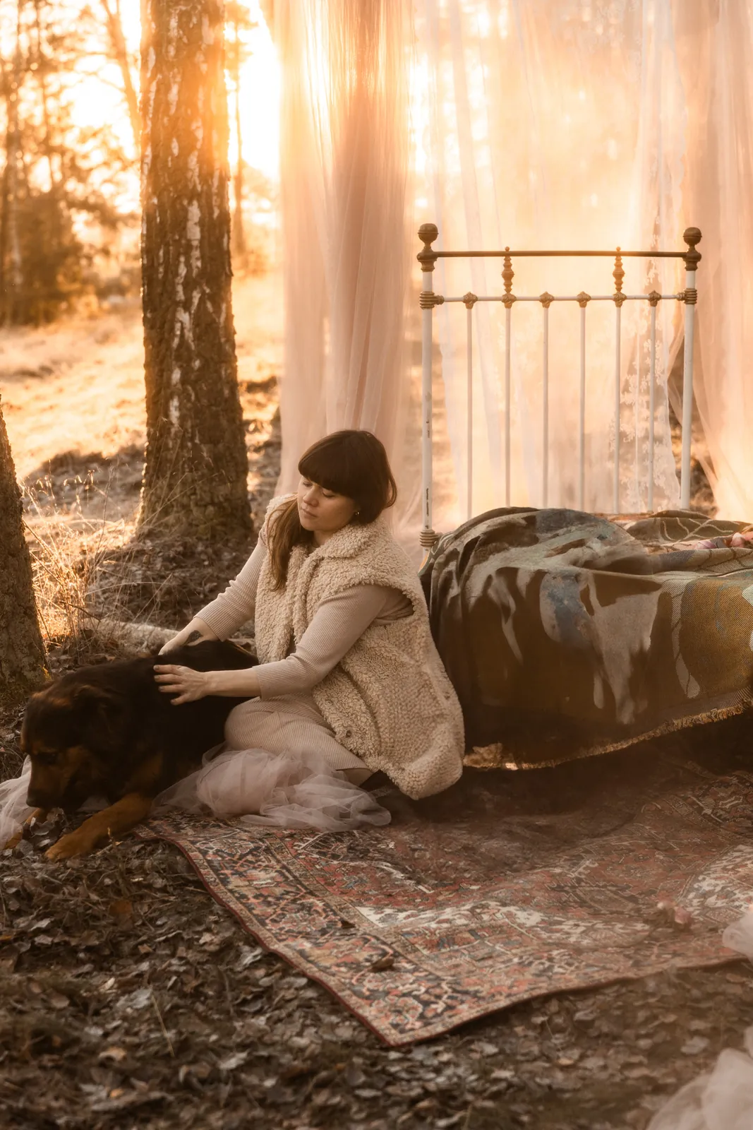 Kobieta w ciąży w naturalnym świetle wśród drzew głaszcze psa. W tle magiczna sceneria plenerowej sesji brzuszkowej.