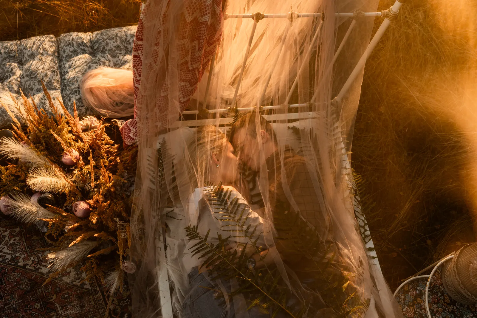 Zakochana para podczas plenerowej sesji zdjęciowej leży na łóżku pośrodku łąki, otula ich delikatny materiał, w tle naturalna kompozycja