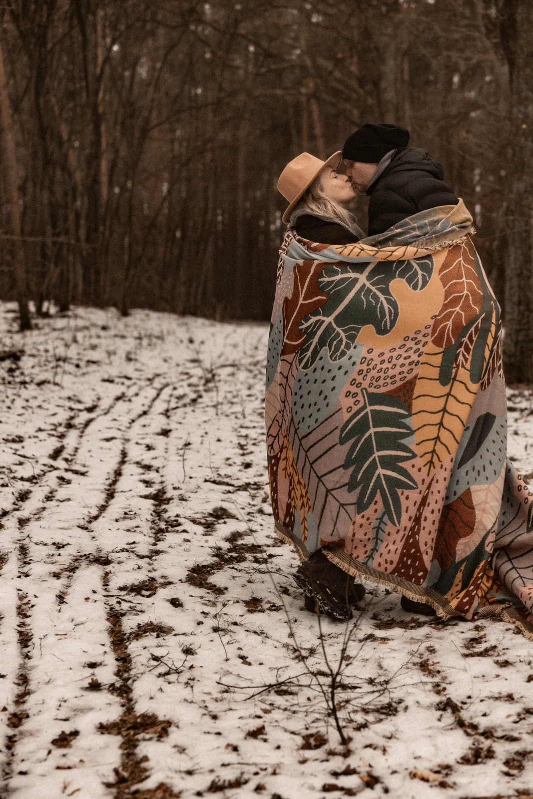 Narzeczeni całują się podczas plenerowej sesji zdjęciowej, okryci stylowym kocem stoją w środku lasu