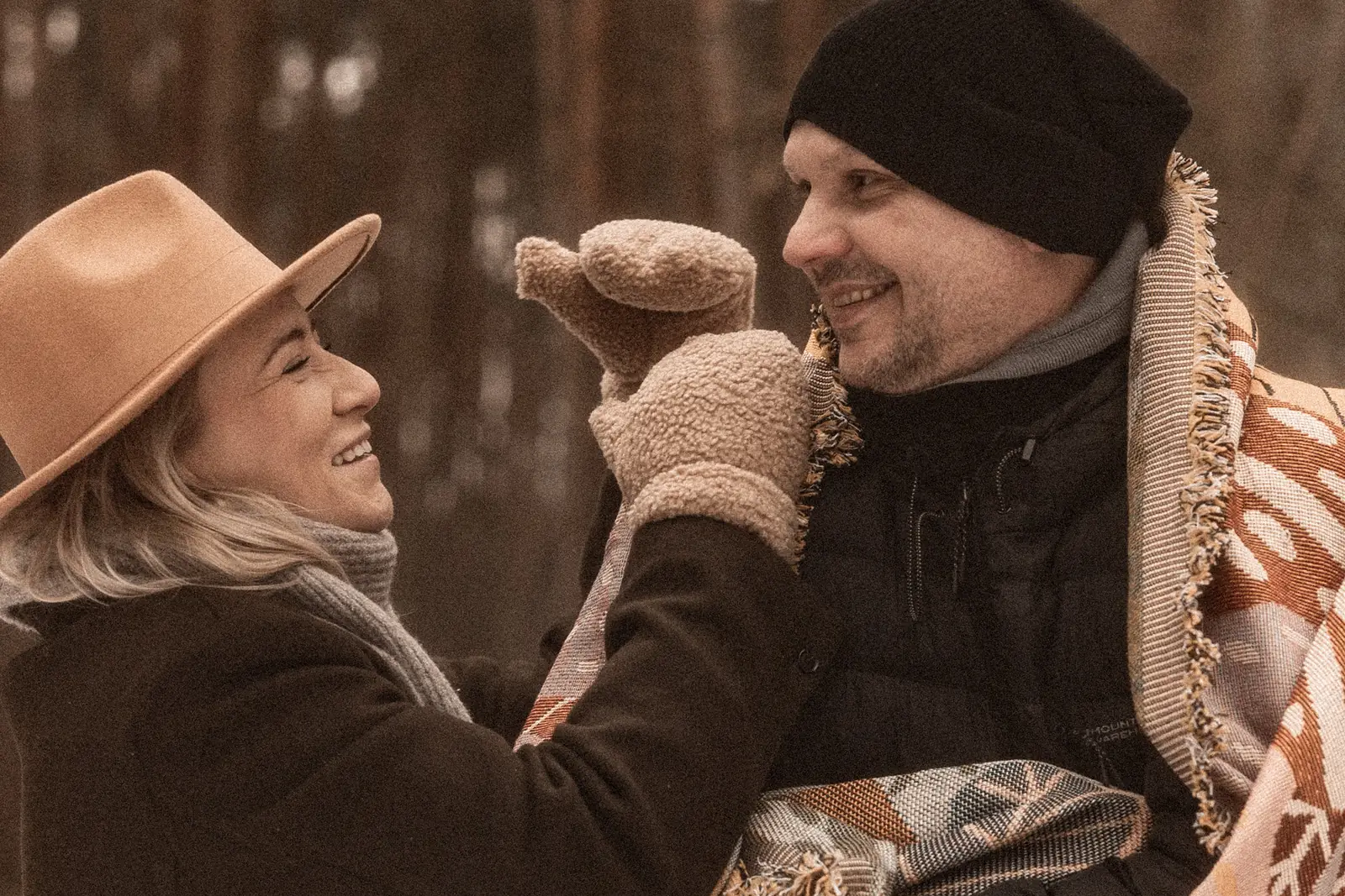 Narzeczeni śmiejący się podczas minimalistycznej sesji zdjęciowej w plenerze, kobieta ma na głowie kapelusz, w tle las i zimowa sceneria