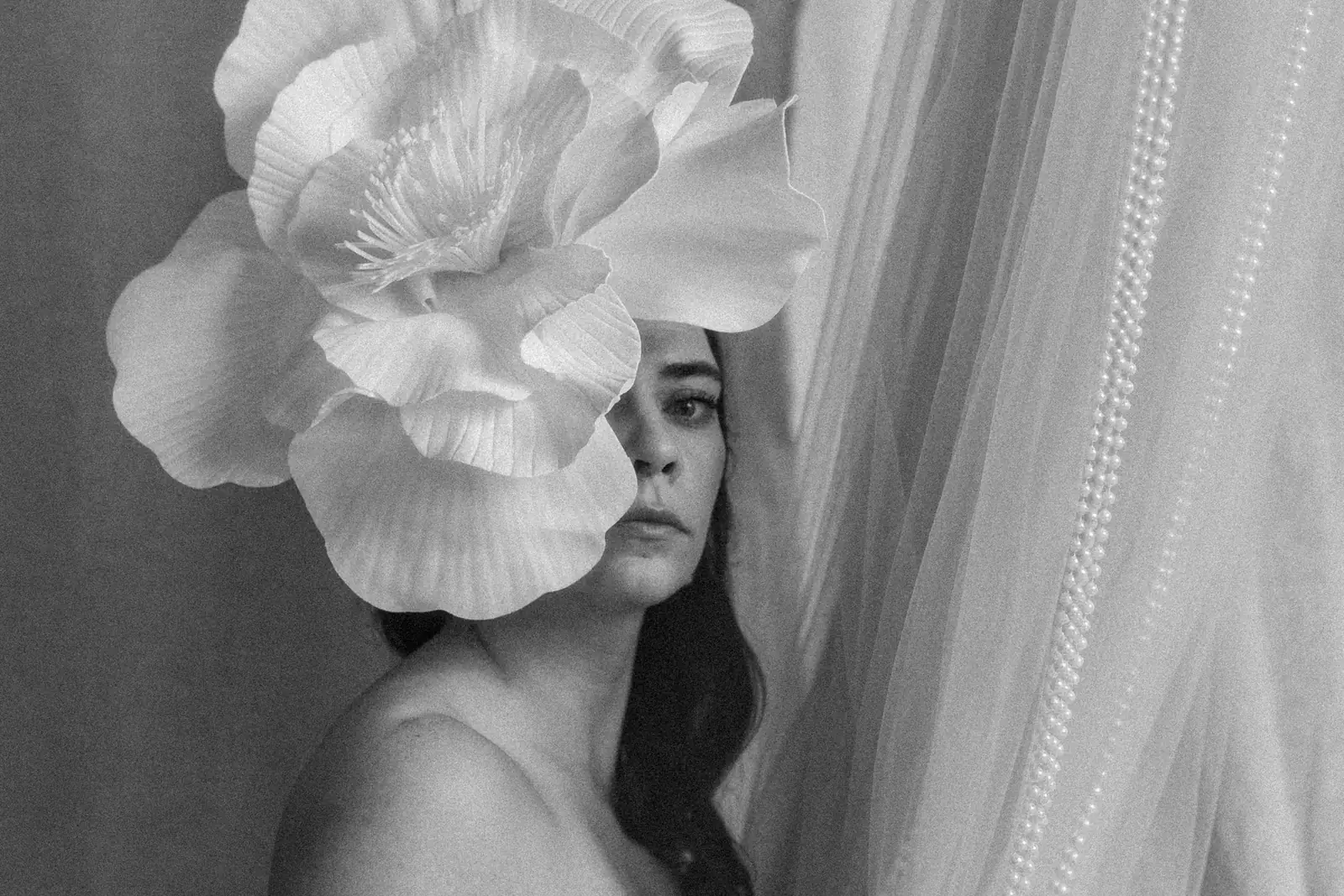 Czarno-biały portret kobiety, duży biały kwiat zdobi kobiece włosy. W tle tiule i białe korale.