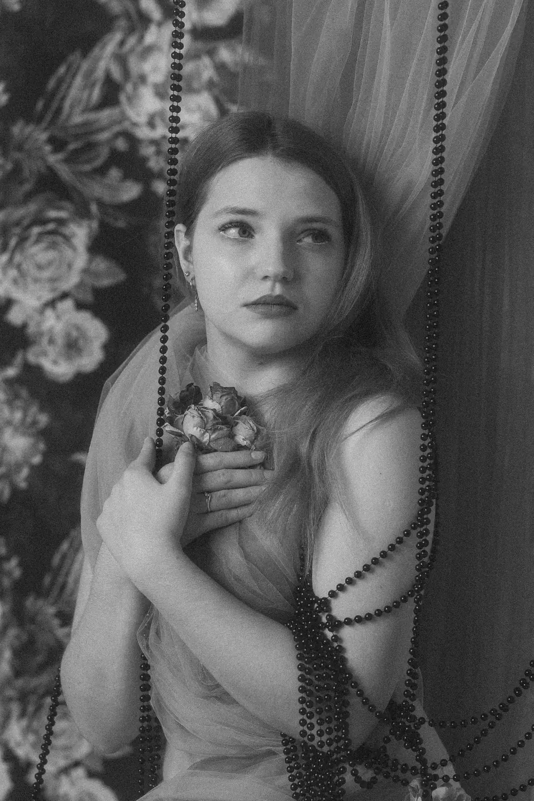 Czarno-biały portret kobiecy. Kobieta trzymająca w ręce kwiat, w tle czarne korale, tło kwiatowe i delikatny materiał.