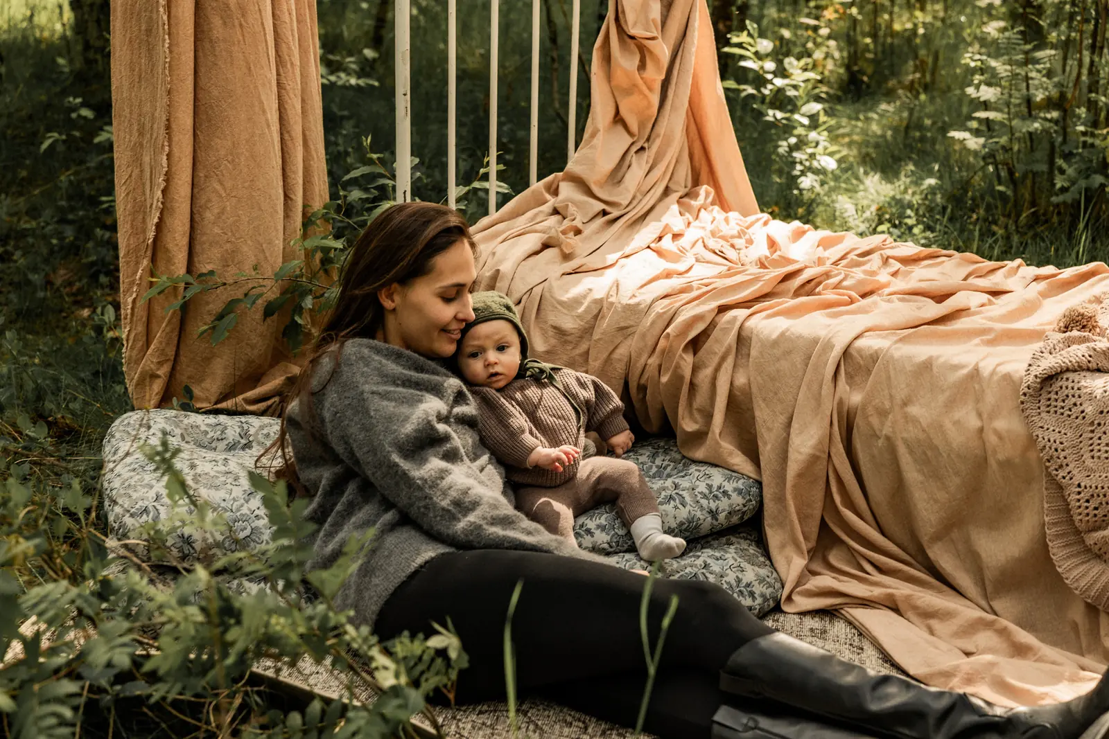 Leśna sesja noworodkowa z łóżkiem ustawionym wśród drzew. Mama obejmuje delikatnie maluszka leżącego na miękkich poduszkach.