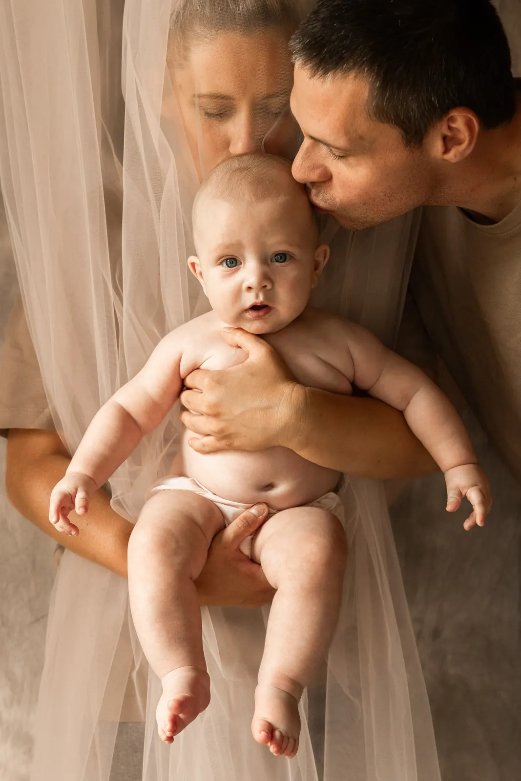 Mama trzymająca niemowlaka na rękach, tata czule całuje główkę dziecka