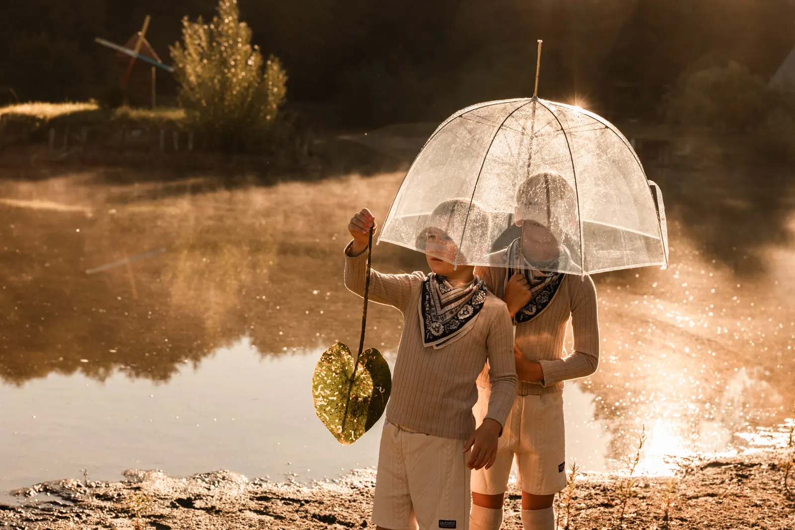 Dzieci nad jeziorem z parasolem, w tle las i wschód słońca