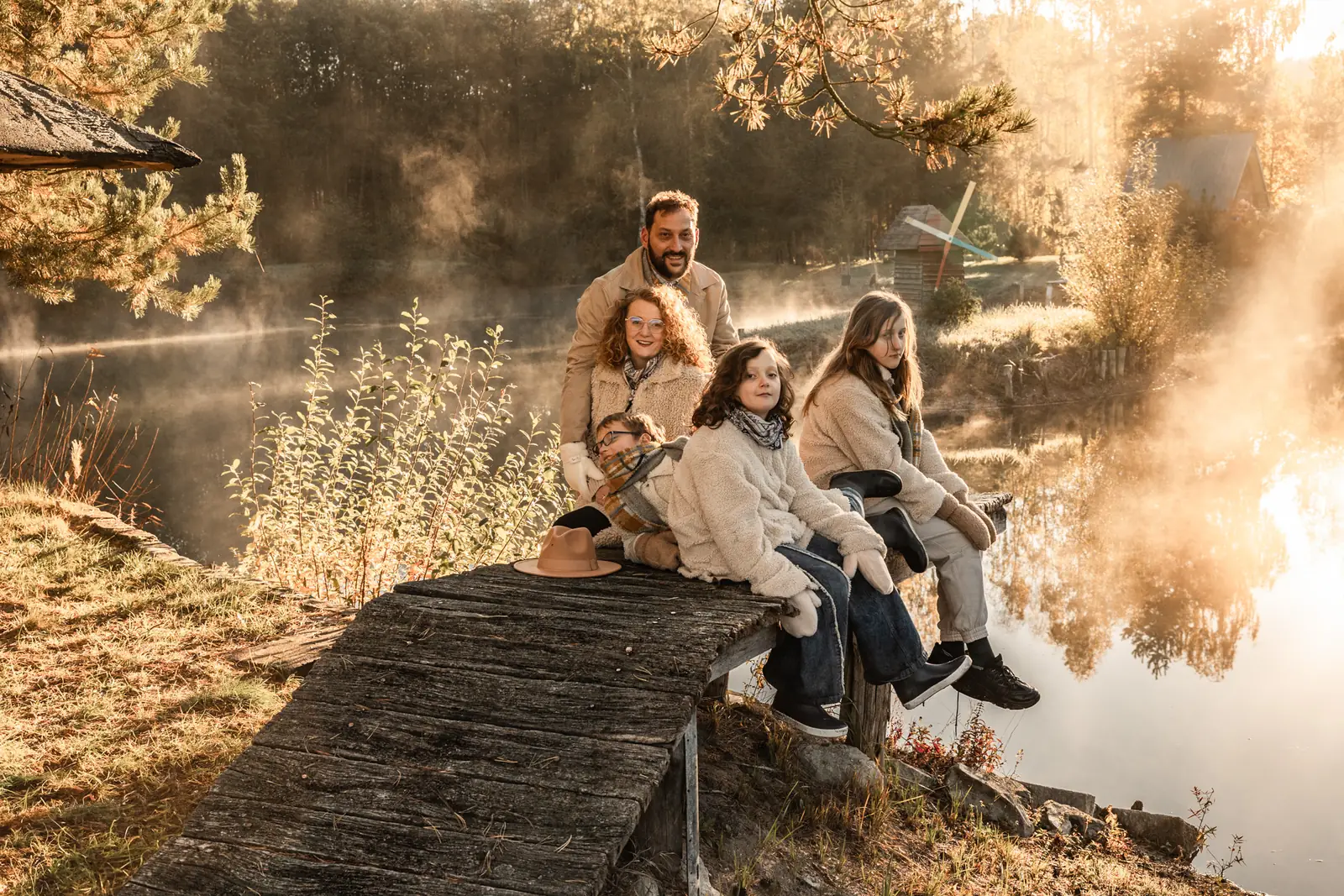 Rodzice z dziecmi siedzą na drewnianym pomoście nad jeziorem. Jesienny plener w mglistym poranku.