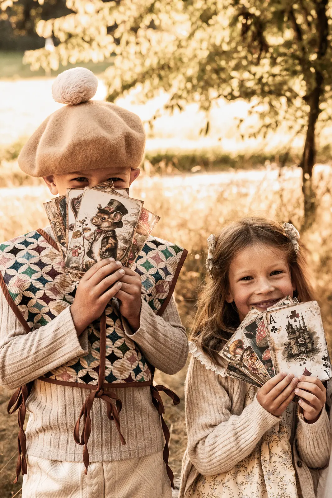 Dzieci bawiące się na łące podczas rodzinnej sesji zdjęciowej. Trzymają w rękach duże karty do gry i spoglądają w obiektyw.
