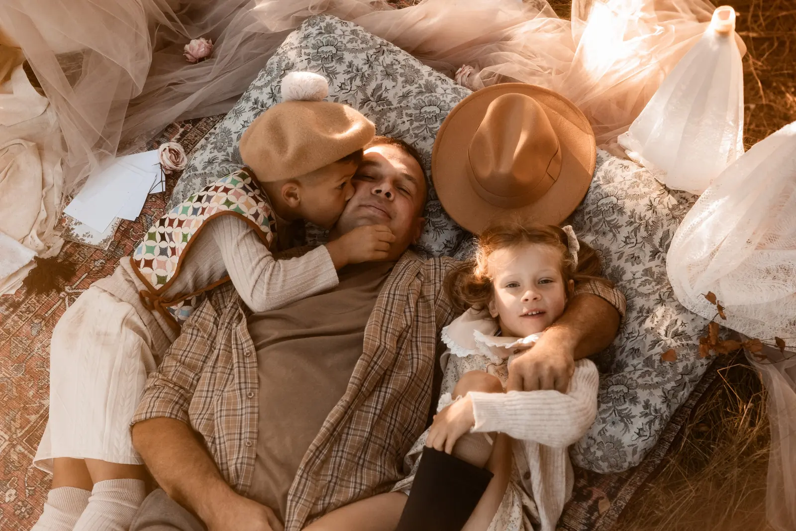 Tata z dziećmi leży na kocu i poduszce w plenerze. W tle kapelusz, kolorowy dywan i miękkie tkaniny zdobiące scenerie.