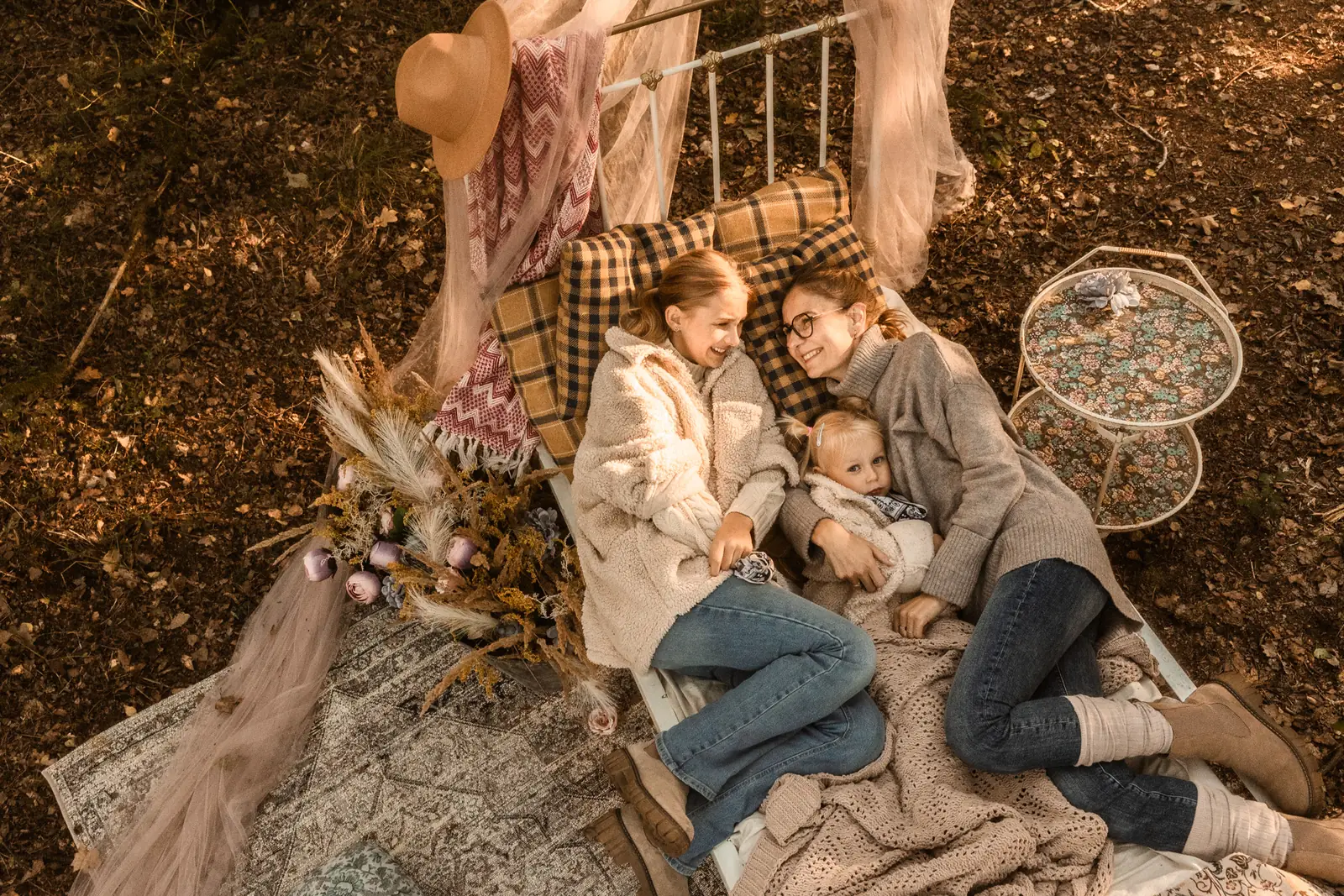 Mama z córkami leży uśmiechnięta na łóżku w środku lasu wśród jesiennych liści.