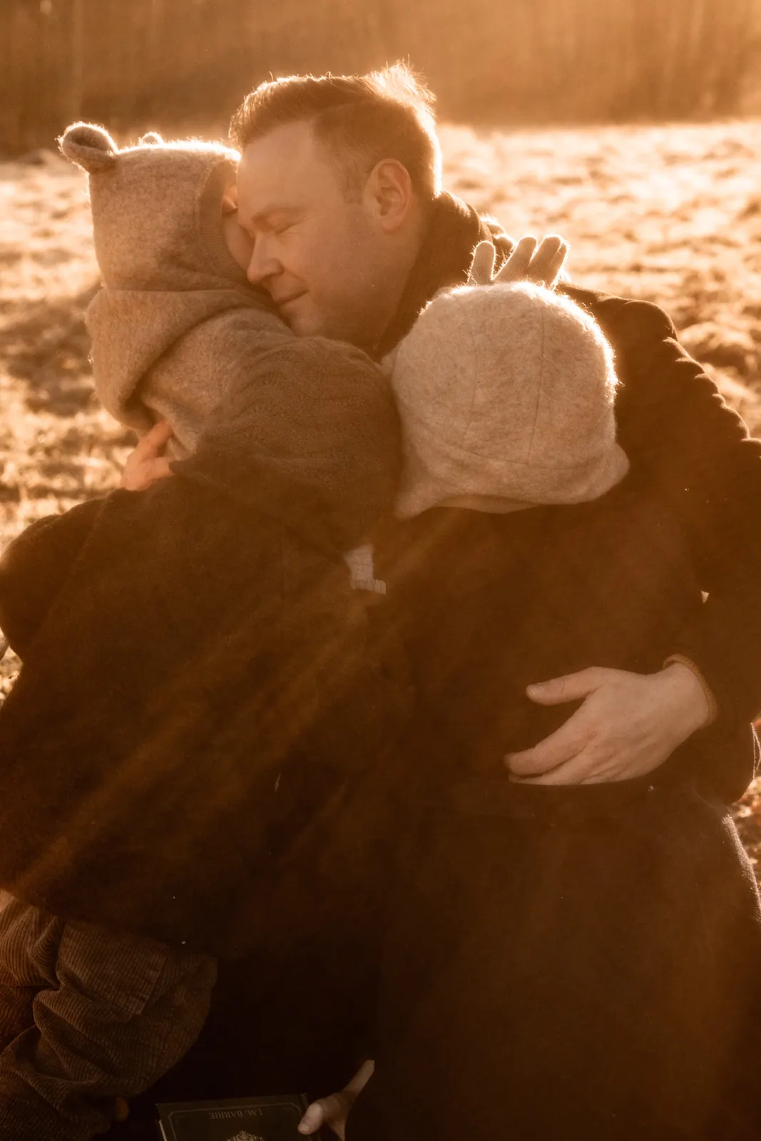 Rodzinna sesja zdjęciowa w ciepłym, naturalnym klimacie - tata przytulający swoje dzieci. W tle zimowa sceneria.