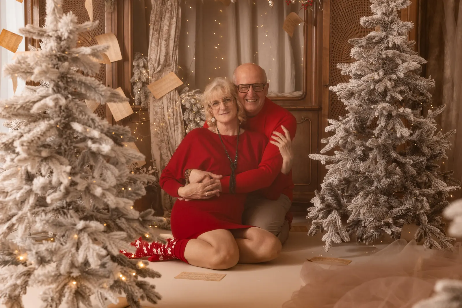 Para w czerwonych swetrach czule przytula się w trakcie świątecznej sesji zdjęciowej, w tle choinki, lampki i modne ozdoby świąteczne