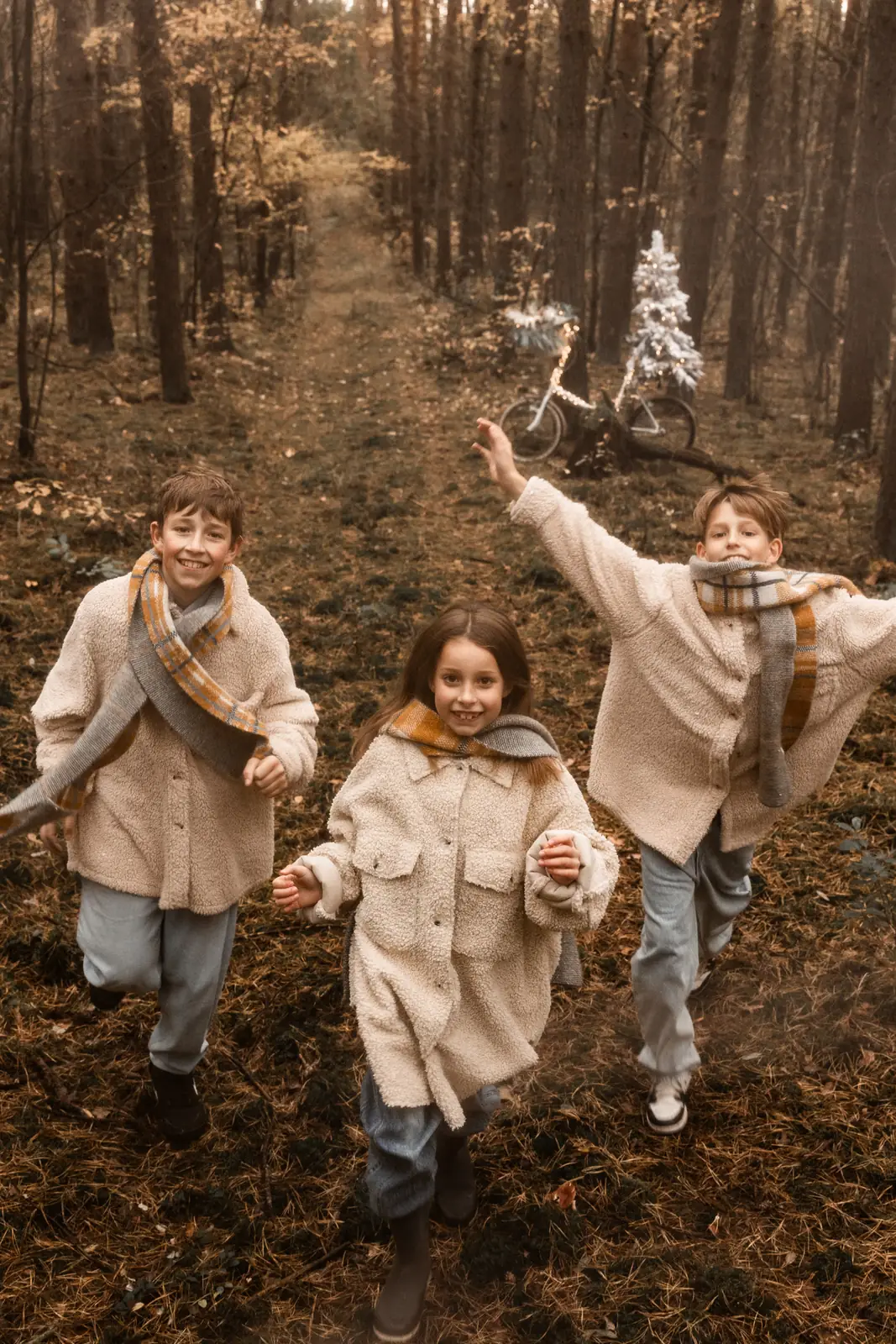 Trójka rodzeństwa biegnie przez las w trakcie świątecznej sesji zdjęciowej, w tle drzewa, rower i choinka