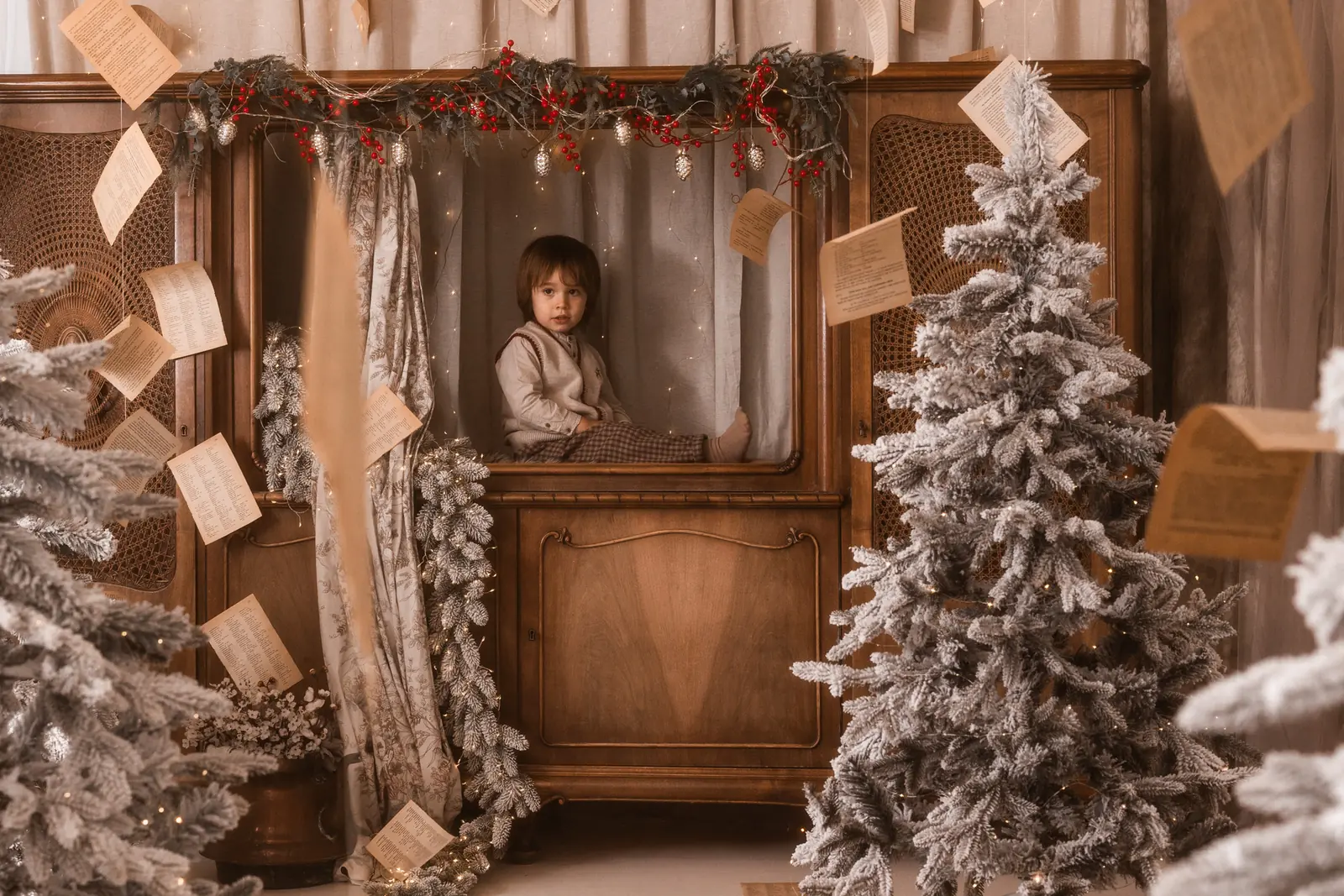 Dziecko siedzi na retro kredensie w studio podczas sesji bożonarodzeniowej. W tle choinki i ozdoby świąteczne.