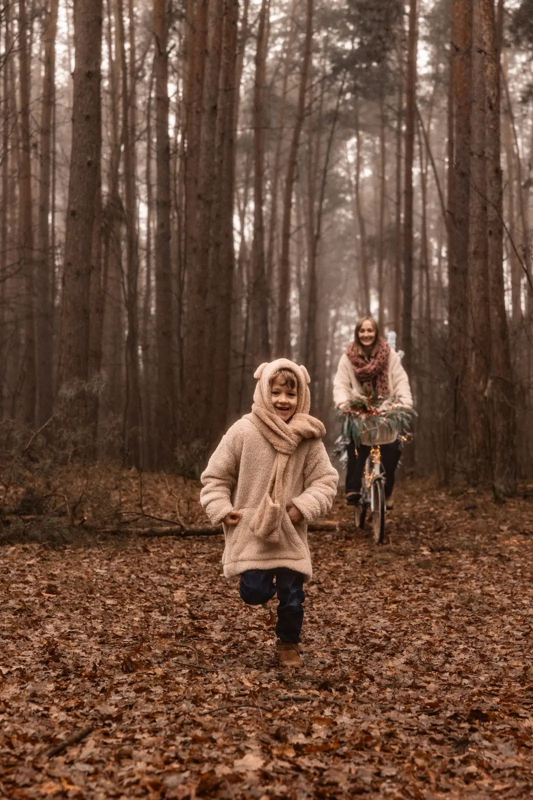 Mama jedzie przez las na rowerze z choinką, a obok niej biegnie uśmiechnięte dziecko.