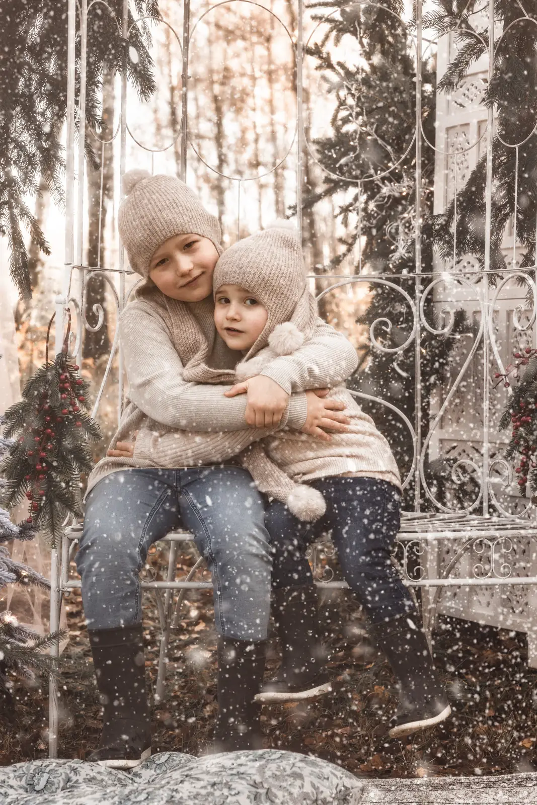 Dzieci siedzą na białej ławeczce przytulone do siebie podczas świątecznej sesji zdjęciowej, w tle śnieg i choinka