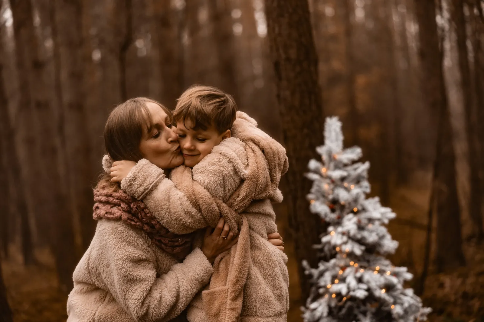 Mama czule przytula dziecko podczas minimalistycznej sesji zimowej w lesie. W tle choinka z świecącymi lampkami.